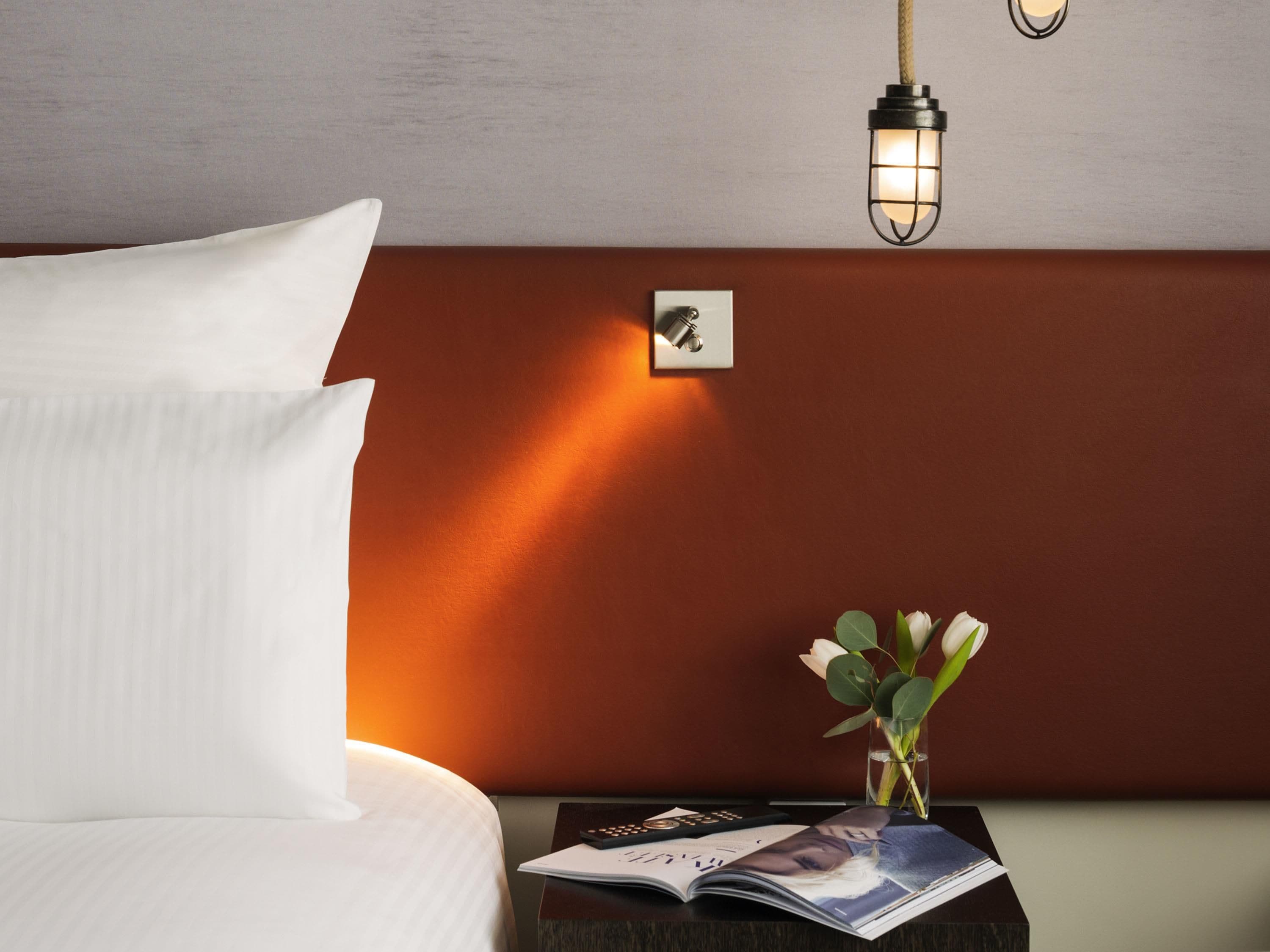 A neatly made bed with white linens, a bedside table with a magazine, a remote control, and a small vase with flowers.
