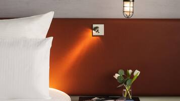 A neatly made bed with white linens, a bedside table with a magazine, a remote control, and a small vase with flowers.