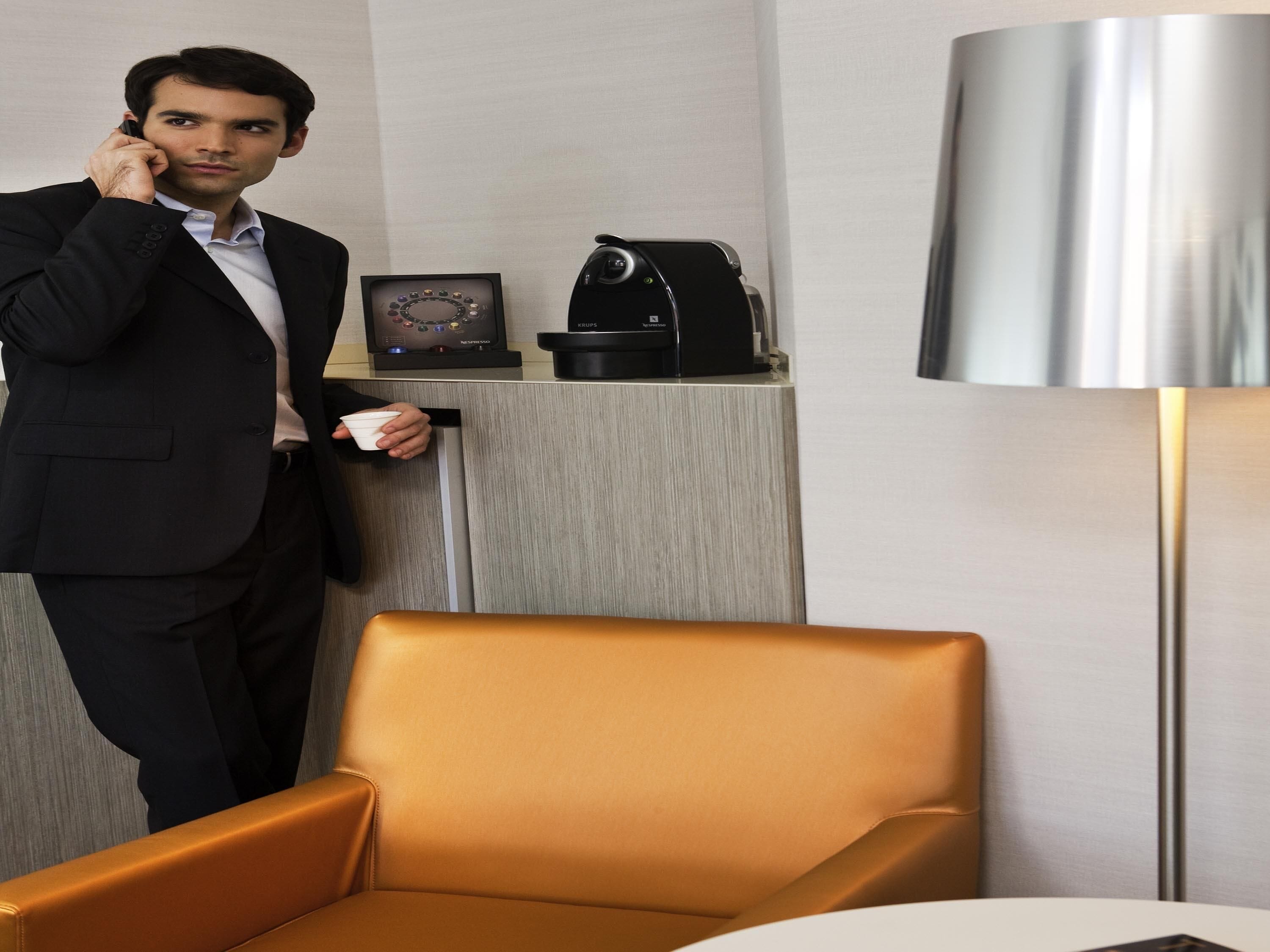 A man in a suit standing by a desk with a lamp, a clock, and a coffee maker.