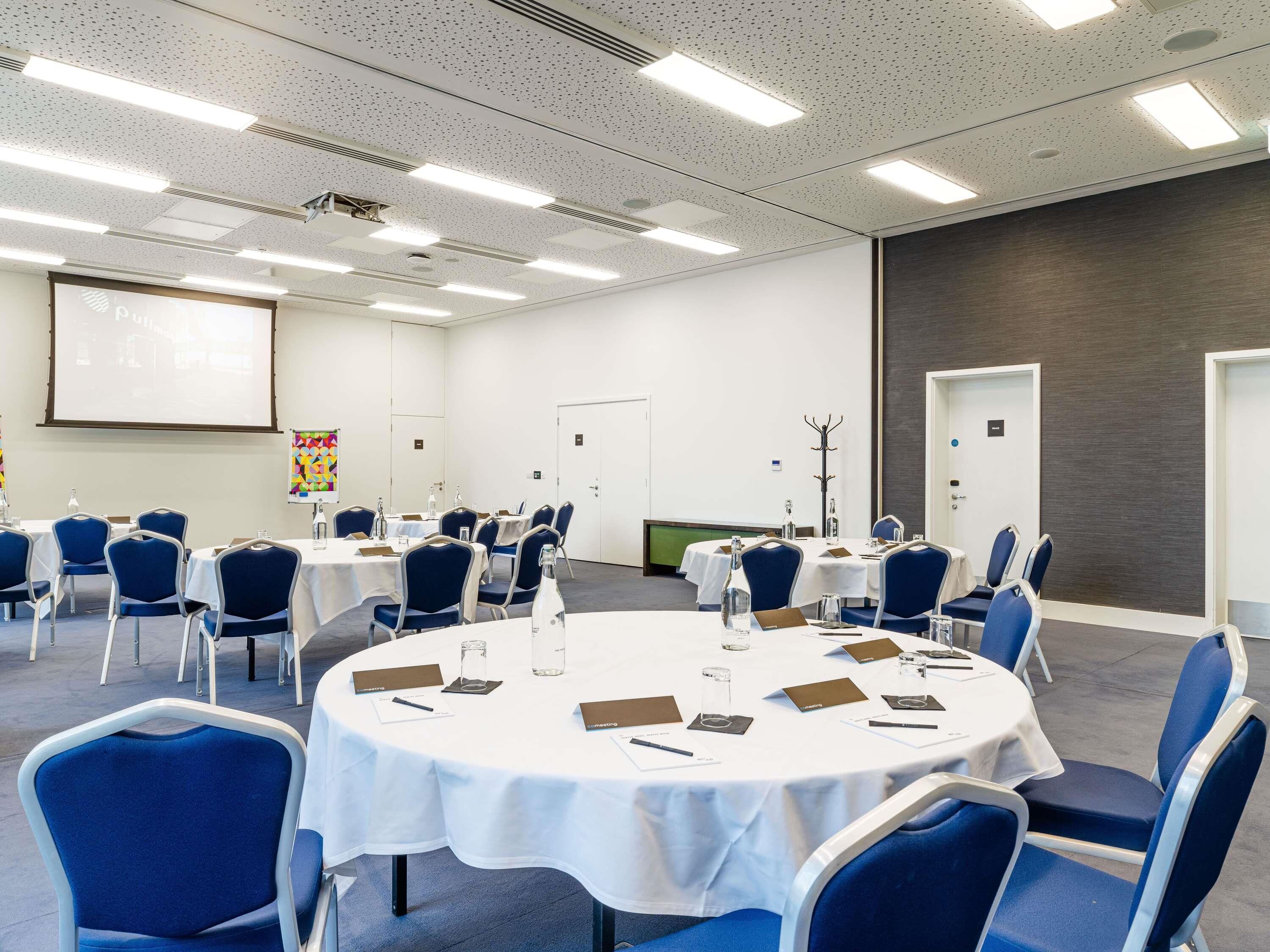 Un salón de conferencias con mesas redondas, sillas azules, manteles blancos y una pantalla para proyector.