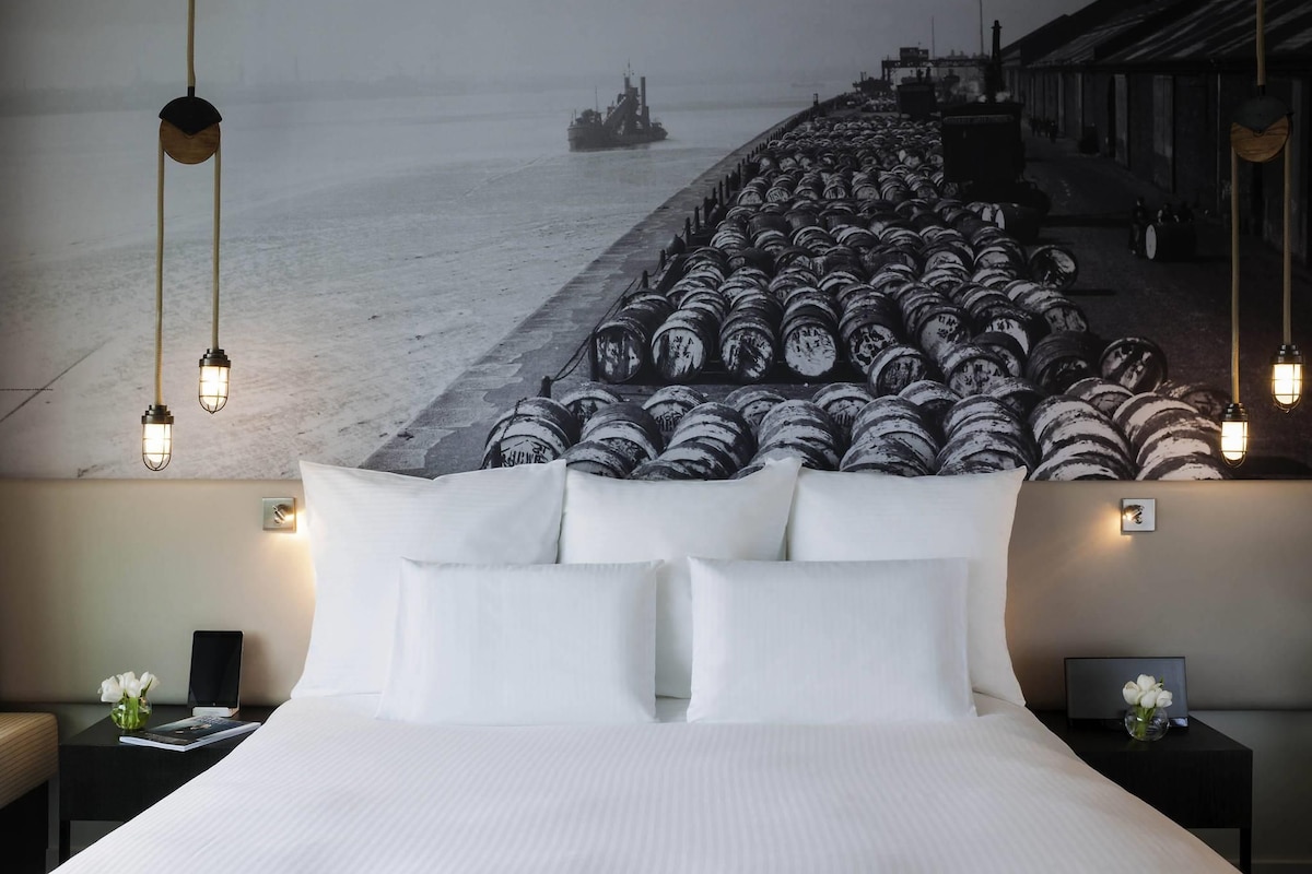 Quarto de hotel com uma cama grande, vista de um navio descarregando barris e design moderno.