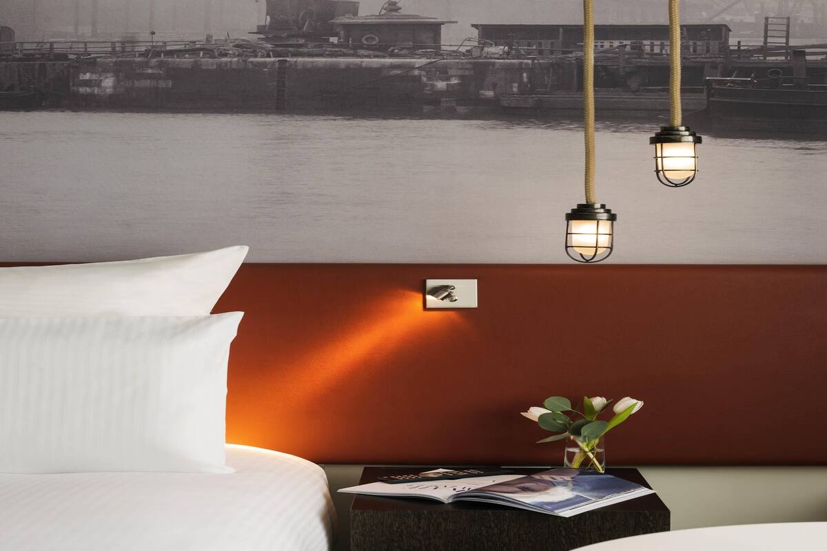 Une chambre d’hôtel avec un lit, une table de chevet sur laquelle repose un magazine, un vase rempli de fleurs et une fresque murale représentant un paysage portuaire.