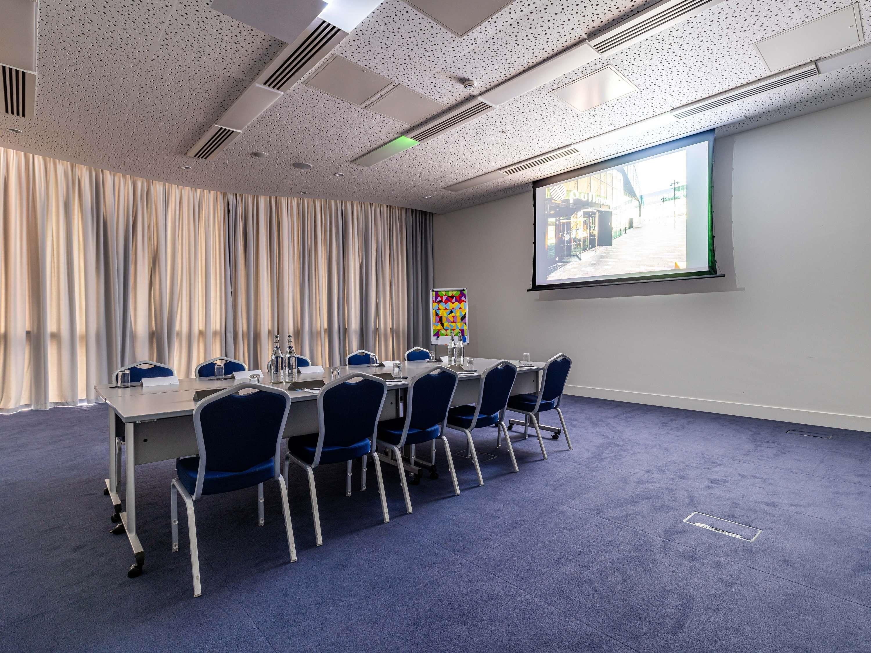 A conference room with a long table, chairs, a projector screen, and a window with curtains.