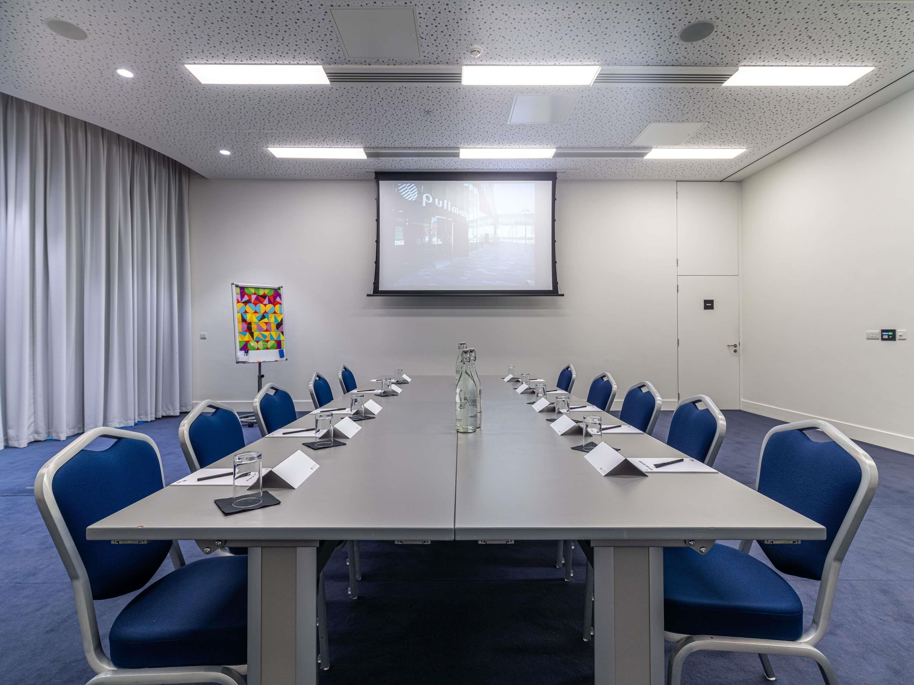 A conference room with a long table, blue chairs, a projector screen, and a whiteboard.