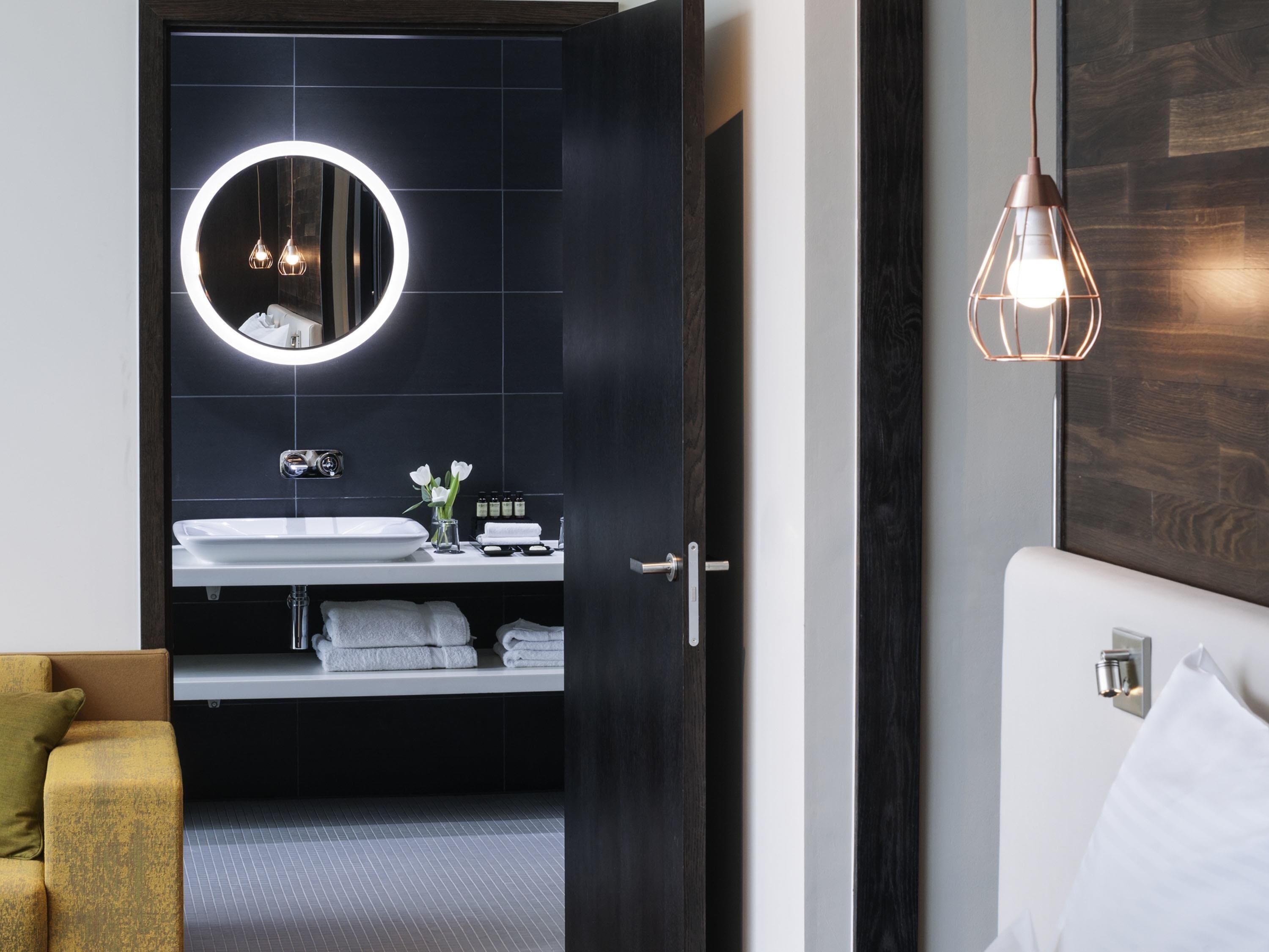 Un baño moderno con un espejo redondo, lavamanos blanco y un perchero.