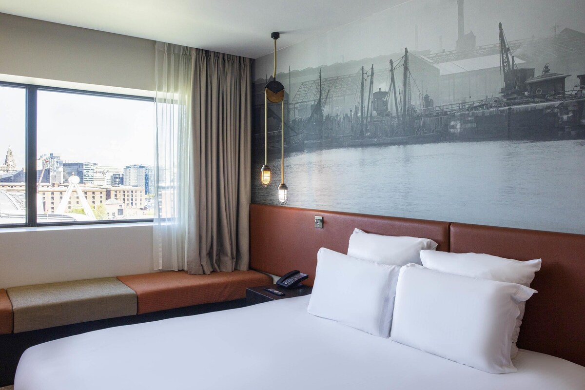Habitación de hotel con una cama grande, vista de un paisaje urbano a través de una ventana, un cuadro colgado en la pared de un puente, y una zona de estar con vistas.