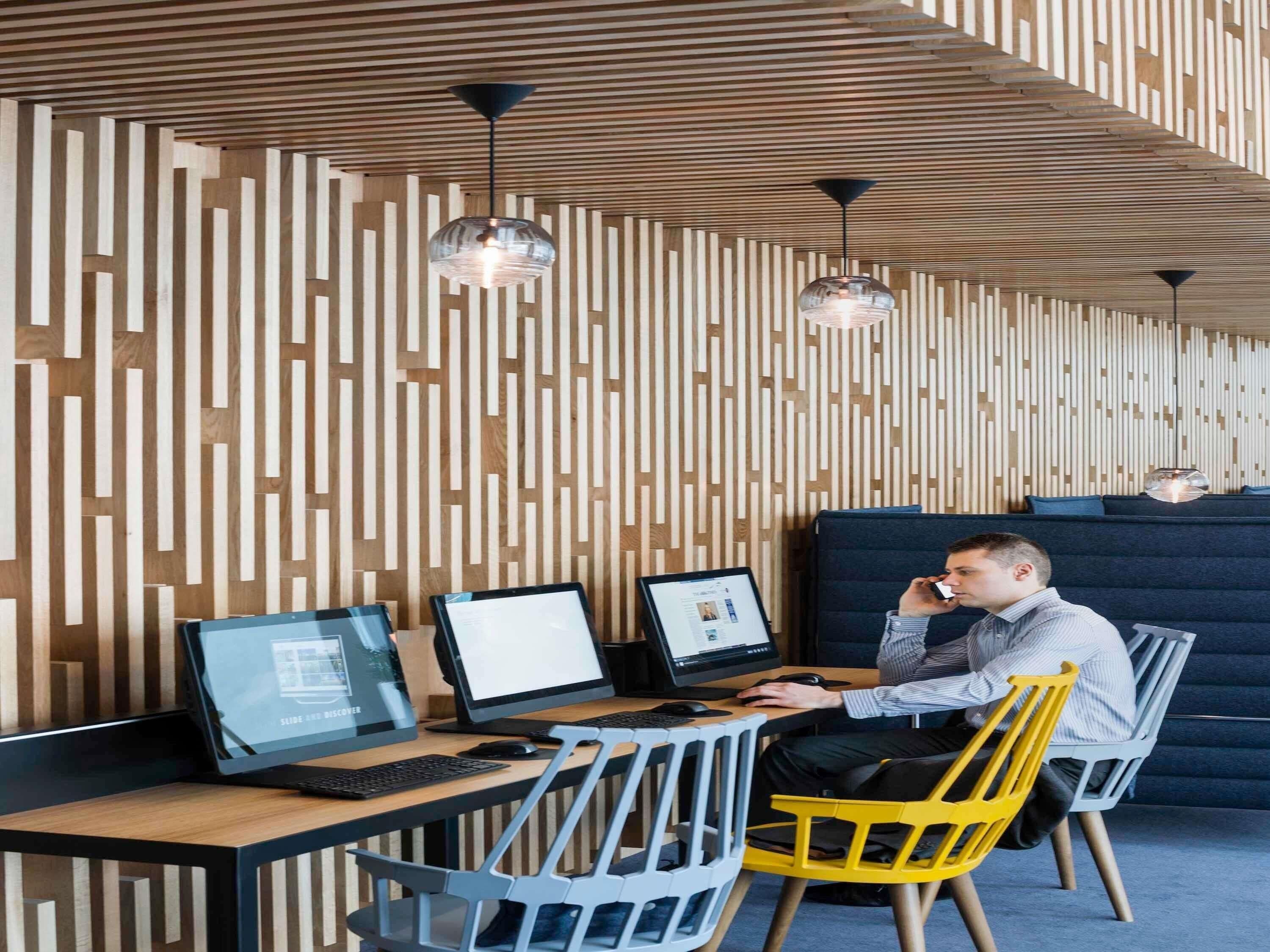 A modern workspace with wooden slats on the wall, two desks with computers, and a person using a mobile device.