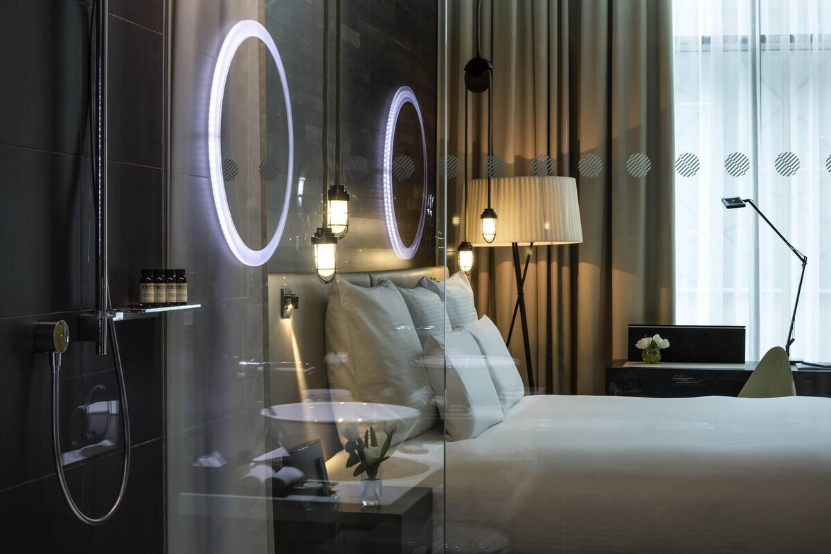 Et moderne hotelværelse med glasvæg omkring brusebad, seng med hvide sengetøj, et natbord med lampe, og udsigt over byen gennem lette gardiner.