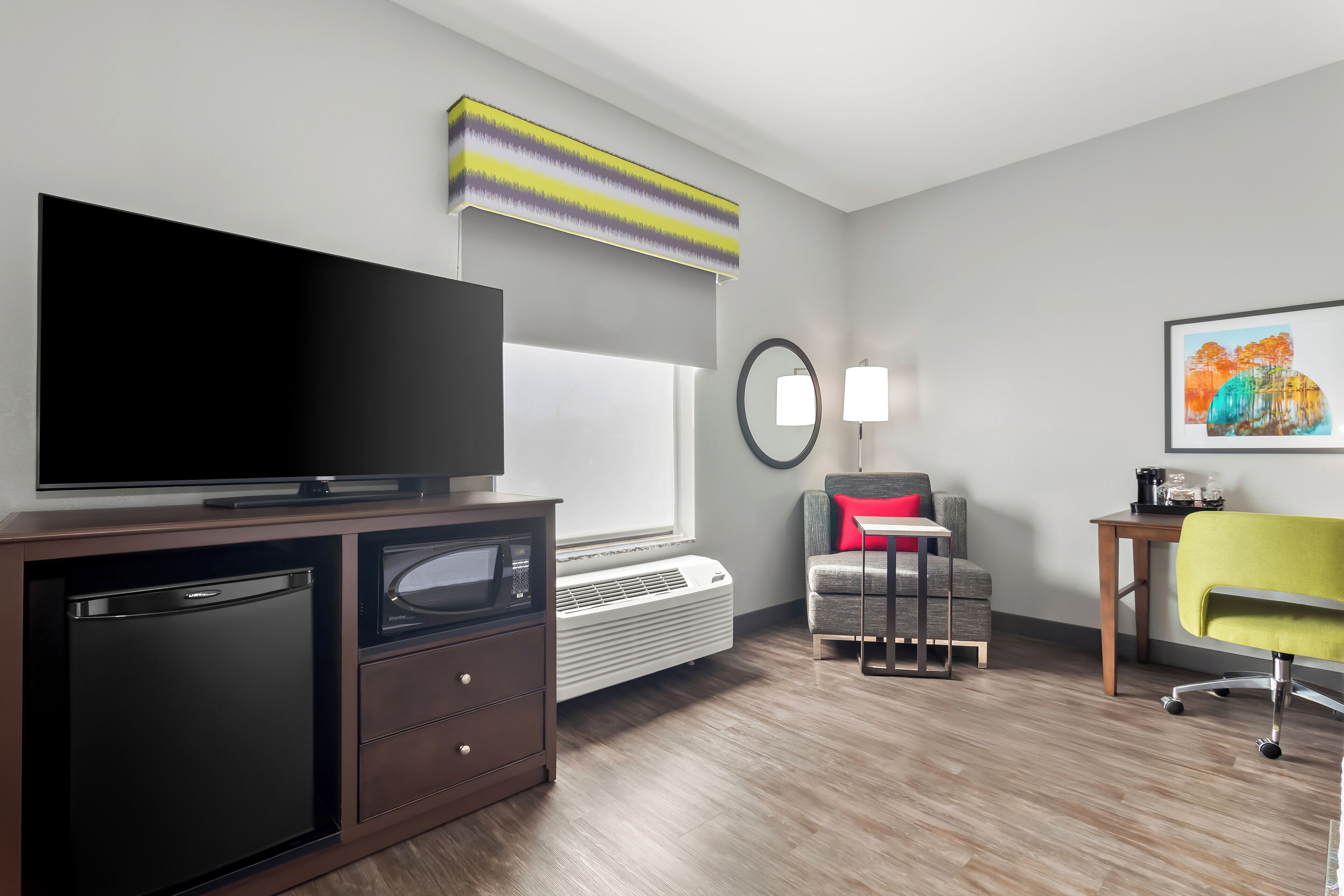 Een moderne hotelkamer met een flatscreen-tv, een minibar, een magnetron, een verwarming, een grijze bank met een rood kussen, een groene bureaustoel, een klein houten tafeltje en een schilderij aan de muur.