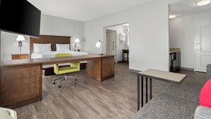 Une chambre d’hôtel avec un lit, un bureau et une chaise, une petite kitchenette et une salle de bain visible à travers une porte ouverte.