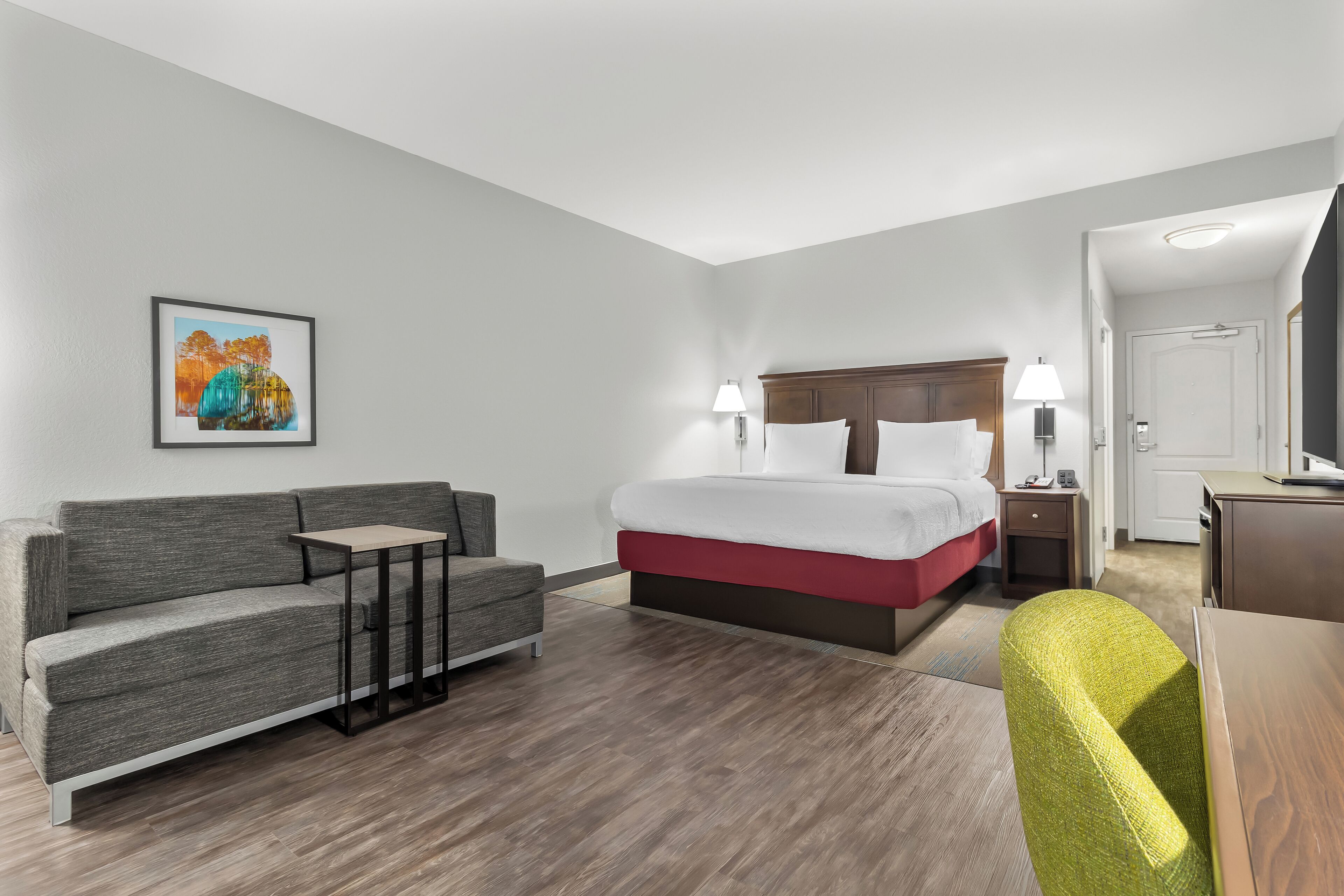 A hotel room with a bed, a sofa, a small table, a lamp, and a TV.