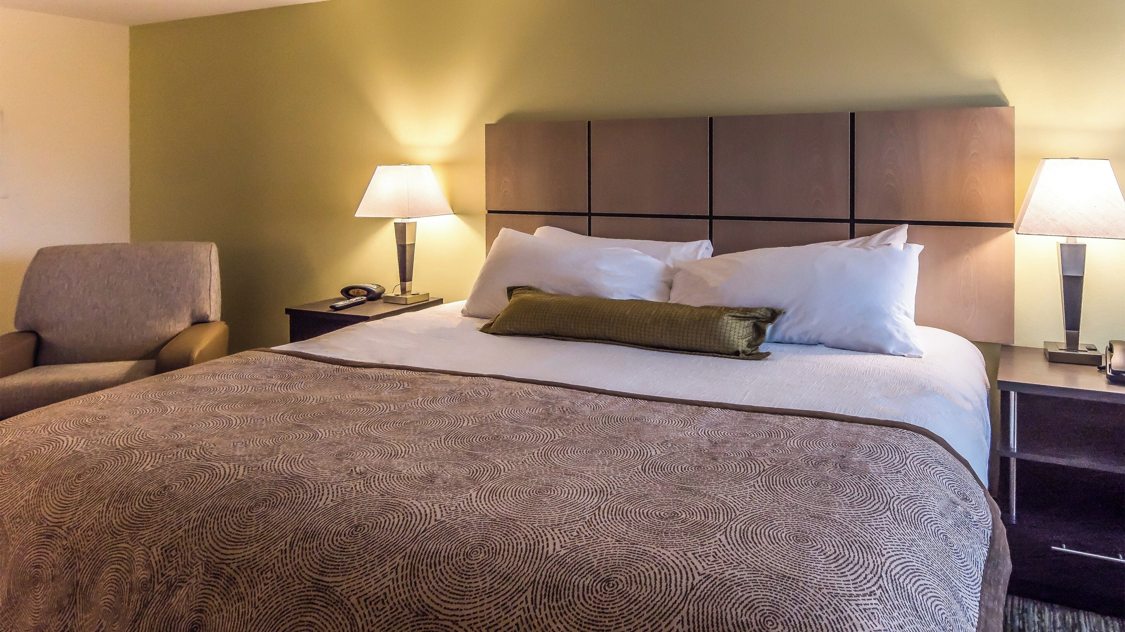 A hotel room with a bed, two bedside lamps, a chair, and a nightstand with a phone and remote control.