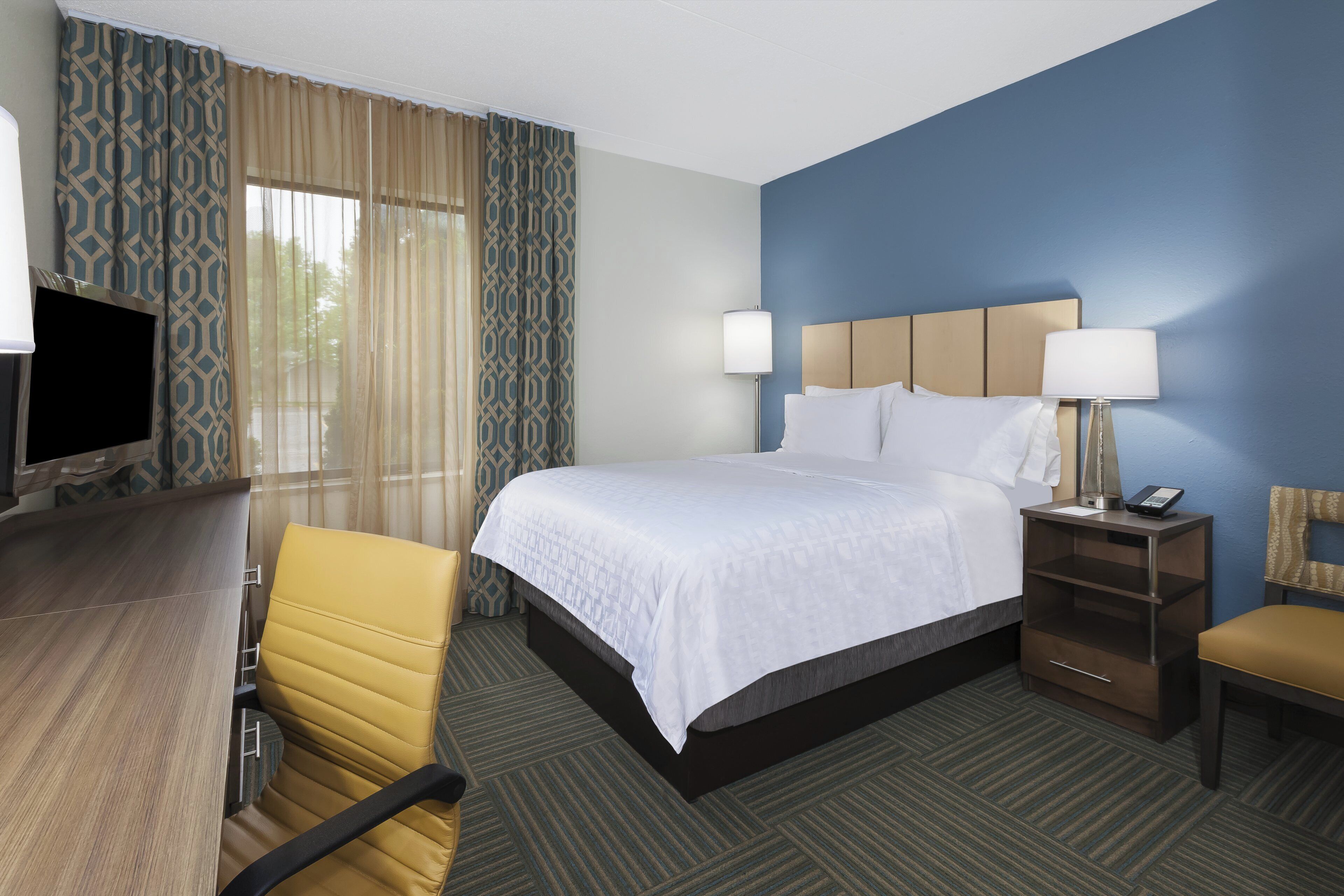 A hotel room with a bed, a desk, a chair, a television, and a nightstand with a lamp and phone.