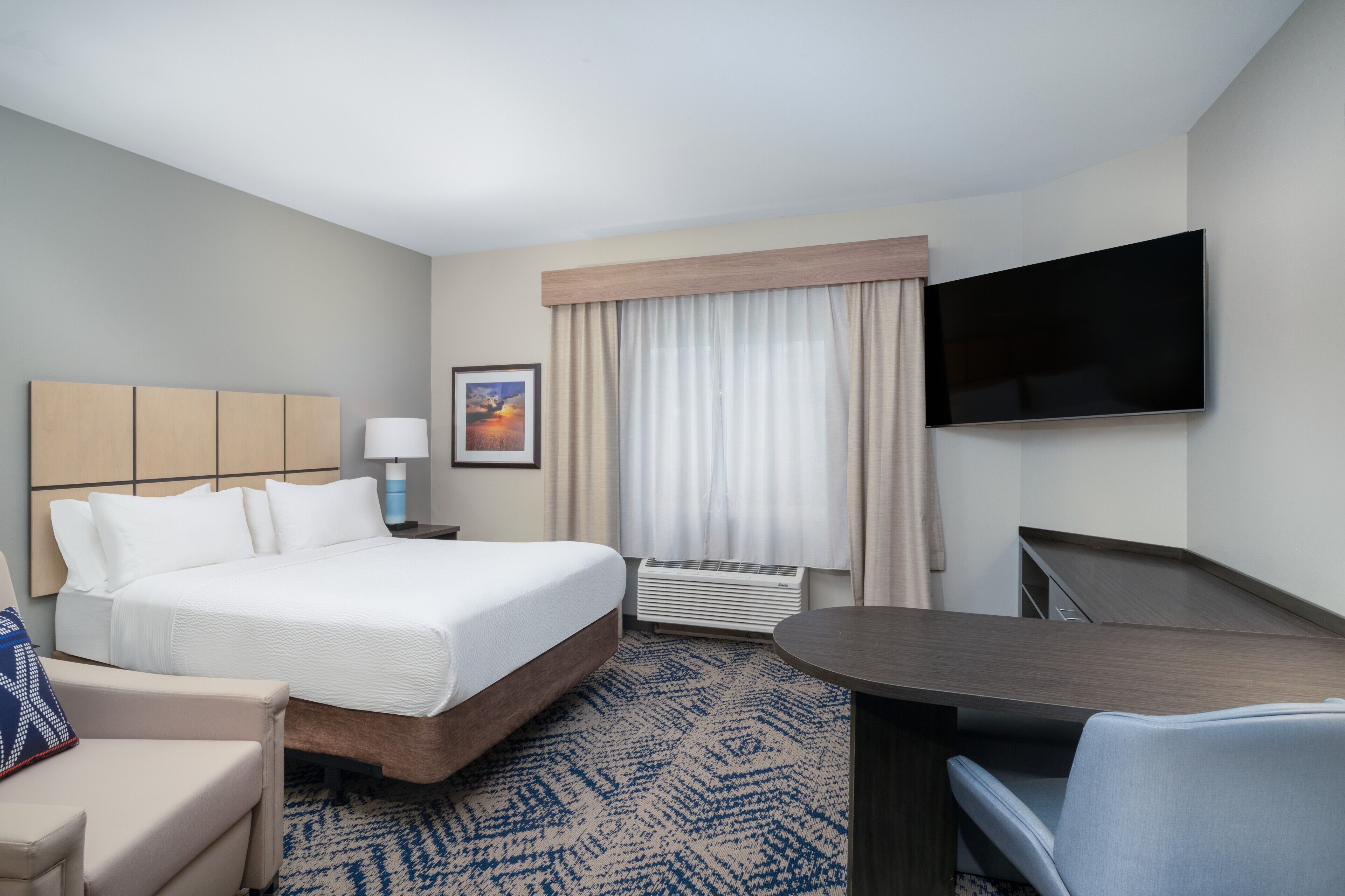 A hotel room with a bed, a desk, a TV, a window with curtains, a lamp, and a framed picture on the wall.