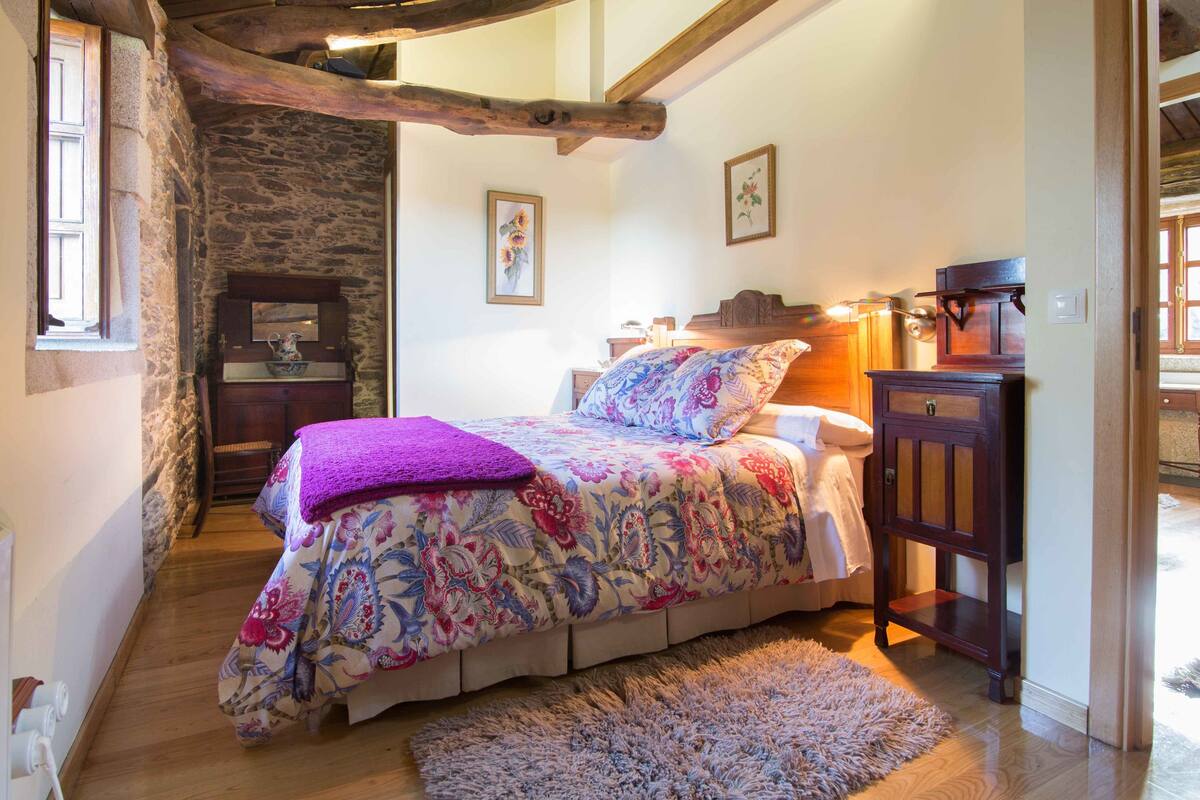 Ein Schlafzimmer mit Bett, Holz-Kopfteil, Nachttisch, Kommode, Steinwand und Fenster.