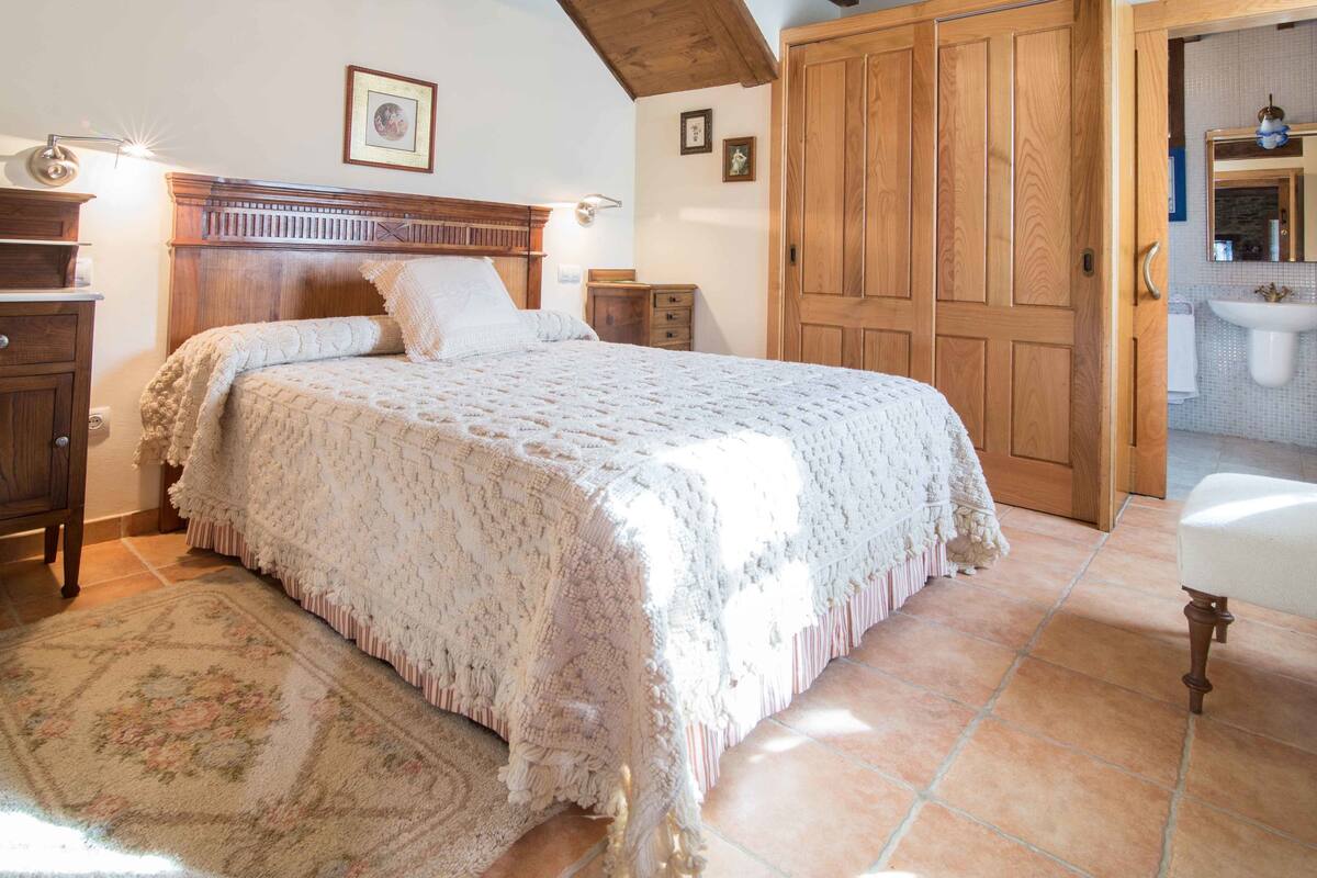 Ein Schlafzimmer mit einem großen Bett, einem Holzkopfende, einem Holzschrank, einem Nachttisch und einem Badezimmer, das durch eine geöffnete Tür sichtbar ist.