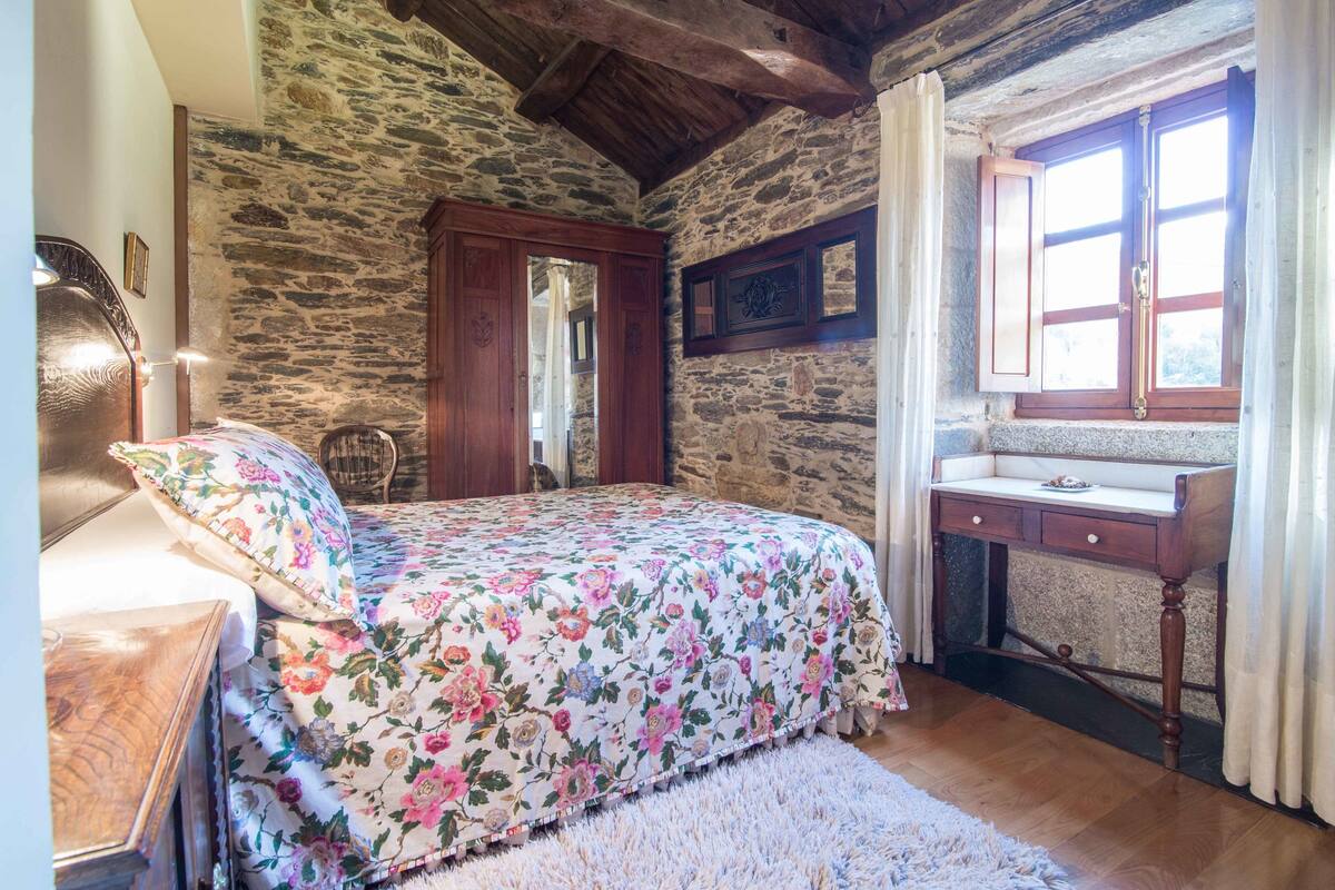Ein Schlafzimmer mit Steinwand, hölzernem Bett, floralem Bettbezug und einem Schreibtisch aus Holz mit Spiegel.