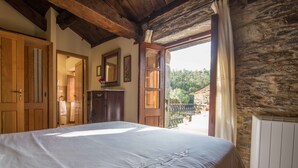 A bedroom with a large bed, wooden ceiling beams, a stone wall, and a view of greenery outside. - Casa Piñeiro (Monfero)