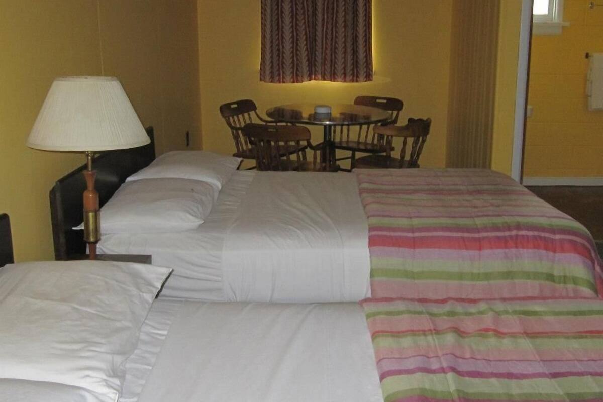A hotel room with a large bed, a bedside table with a lamp, a small round table with chairs, and a window with curtains.