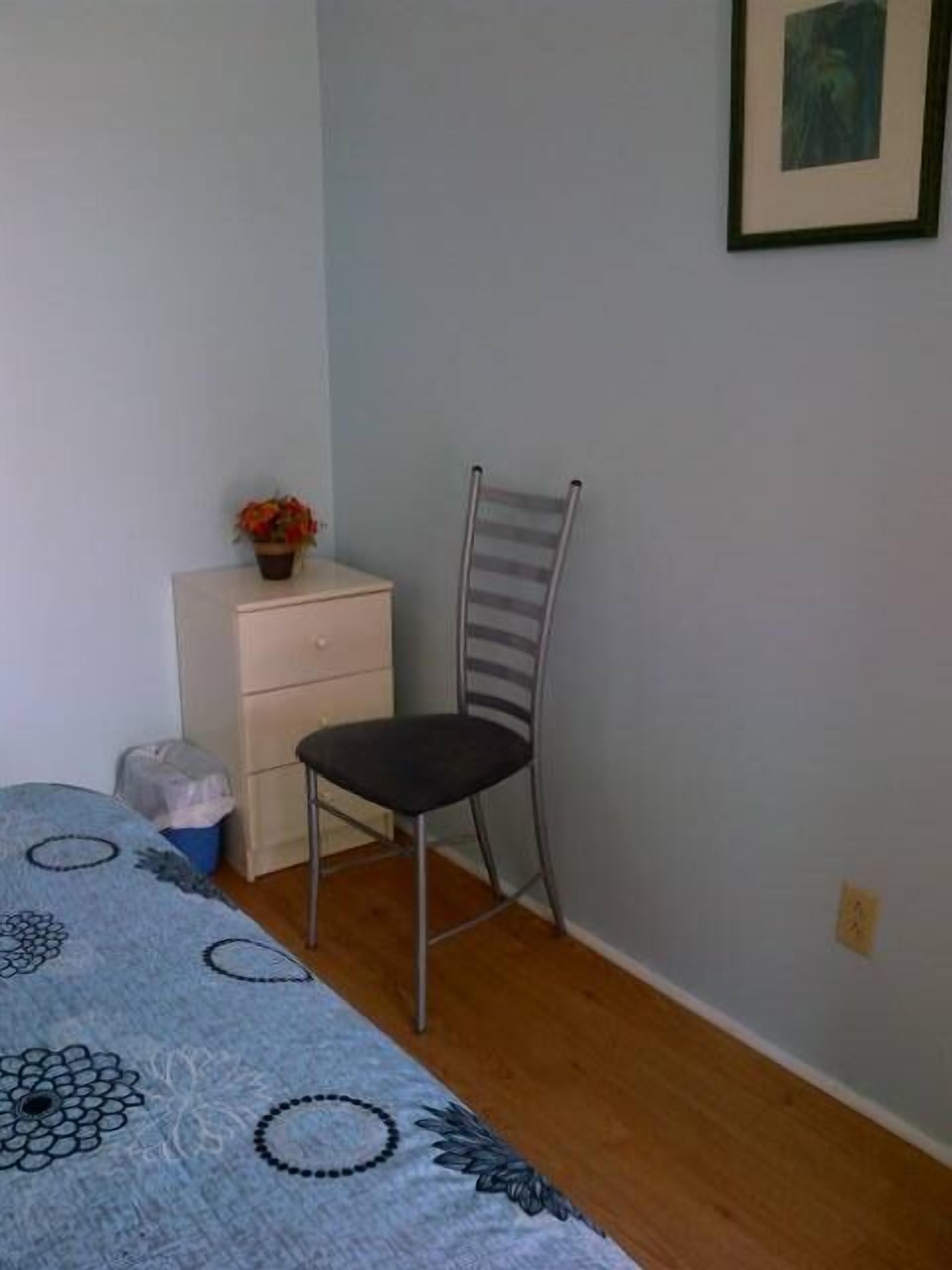 A small, simple room with a bed, a chair, a small table with a plant, and a wall-mounted picture.