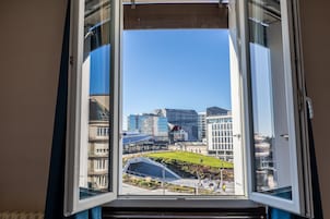 Une vue à travers une fenêtre ouverte sur un ensemble d’immeubles modernes aux façades vitrées et à la pelouse verdoyante.
