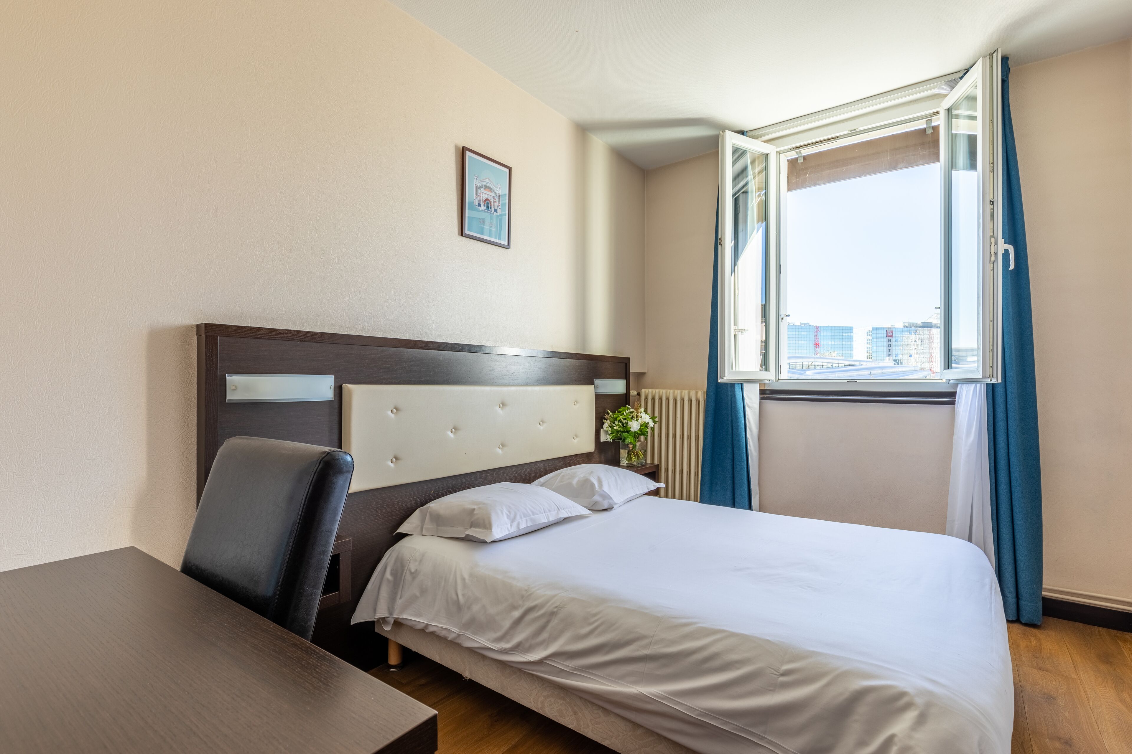 A hotel room with a bed, a desk, a chair, a window with curtains, and a picture on the wall.