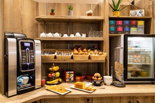 Un buffet de petit-déjeuner comprenant une machine à café, du pain, des viennoiseries, des fruits et diverses boissons.