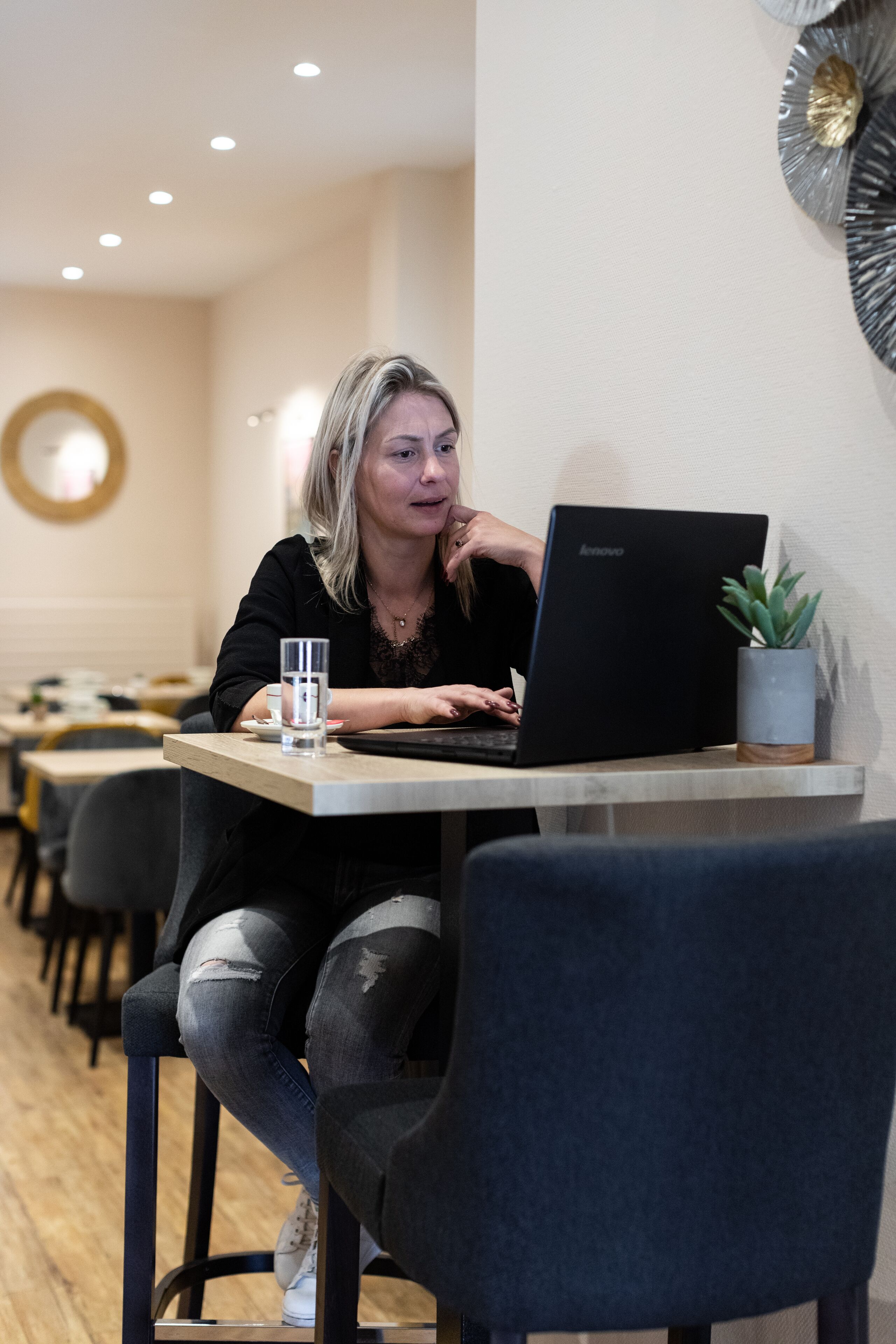 Een persoon zit aan een tafel met een laptop, een glas water en een bloempot.