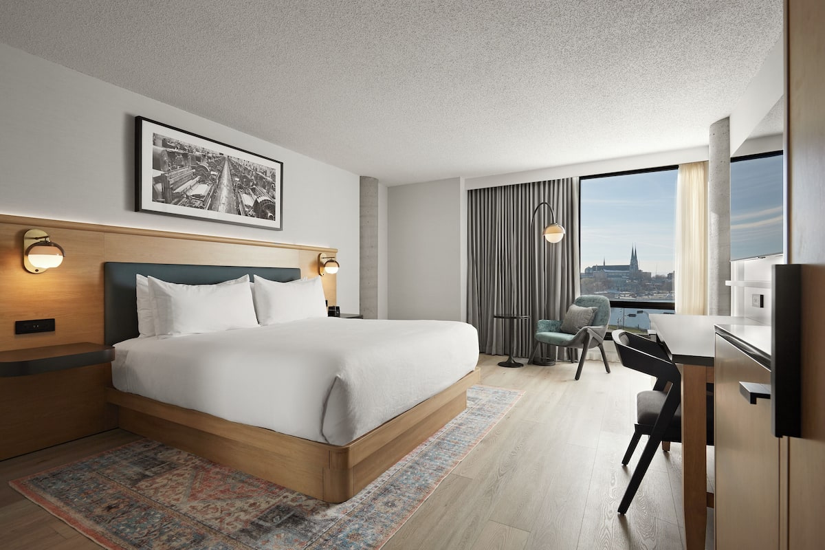 A modern hotel room with a large bed, a desk, and a view of a cityscape through the window.