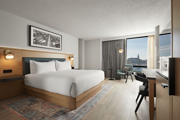 A modern hotel room with a large bed, a desk, and a view of a cityscape through the window. - Hôtel MOCO Valleyfield (Salaberry De Valleyfield)
