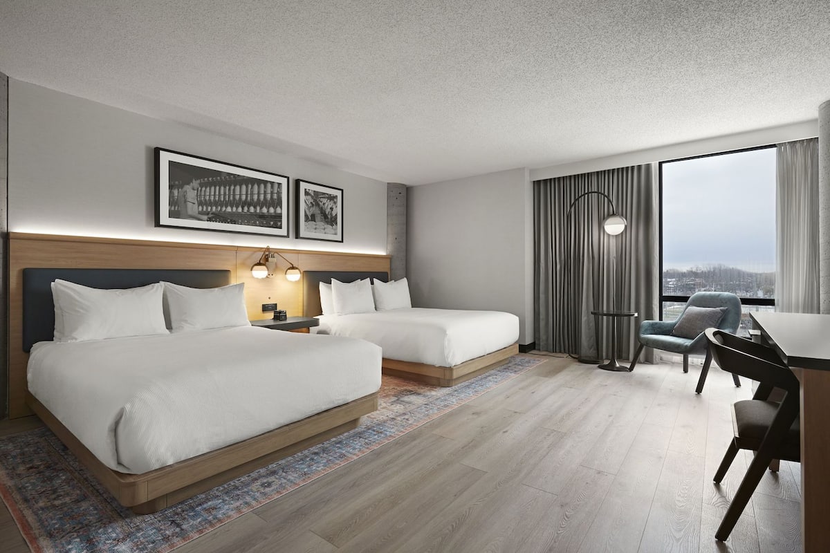 A hotel room with two beds, a desk, a chair, a large window with curtains, and framed pictures on the wall.
