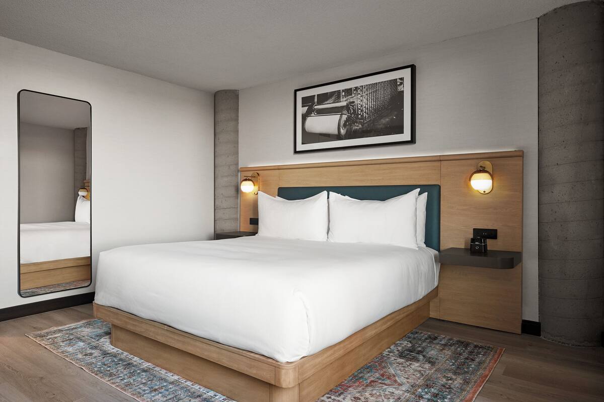 A modern hotel room with a large bed, a bedside table with a lamp, a wall-mounted picture, and a large mirror.