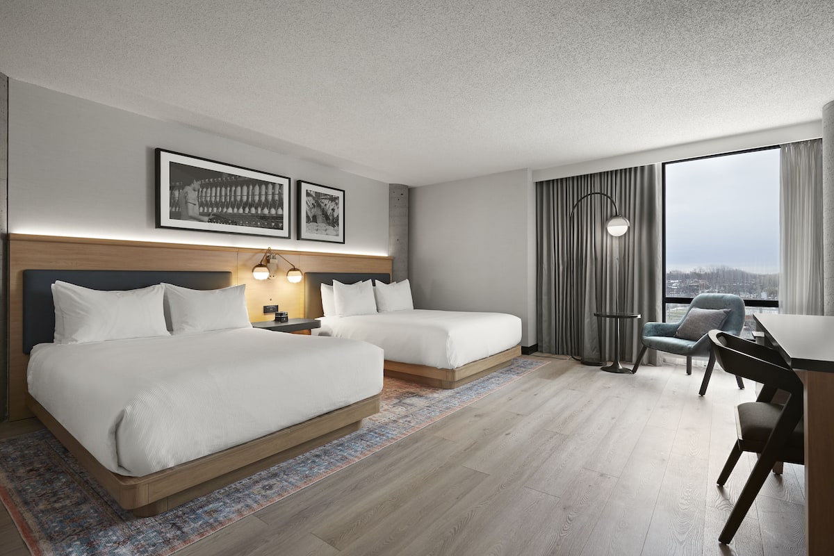 A modern hotel room with two beds, a desk, and a large window with a view.