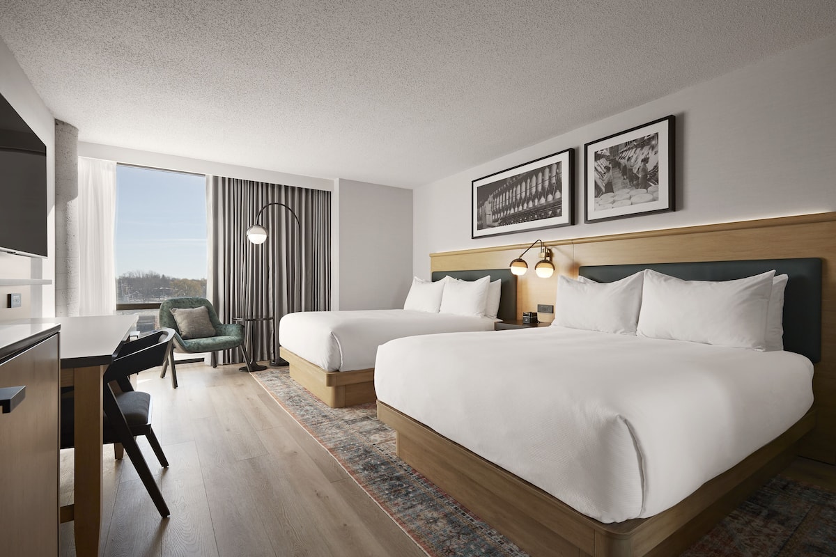 A hotel room with two beds, a desk, a chair, and a large window.