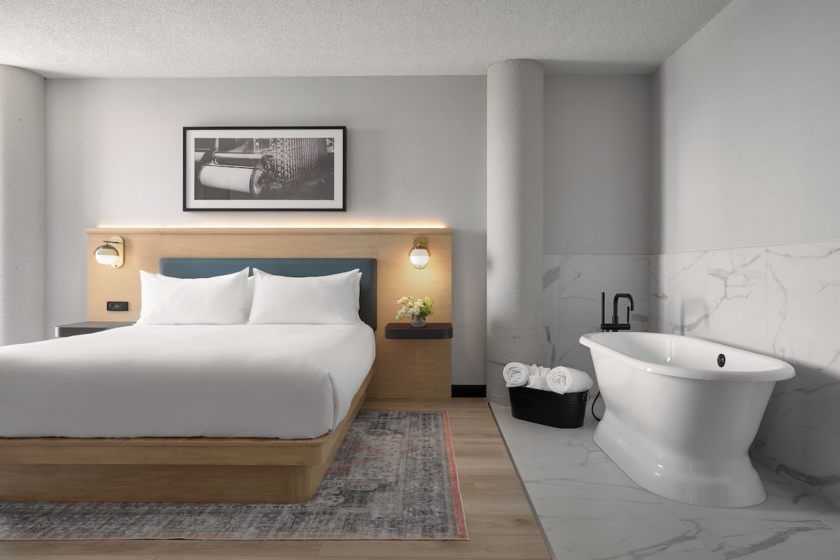 A modern bedroom with a bed, bedside tables, a nightstand, a wall-mounted light, a framed picture, and a freestanding bathtub.
