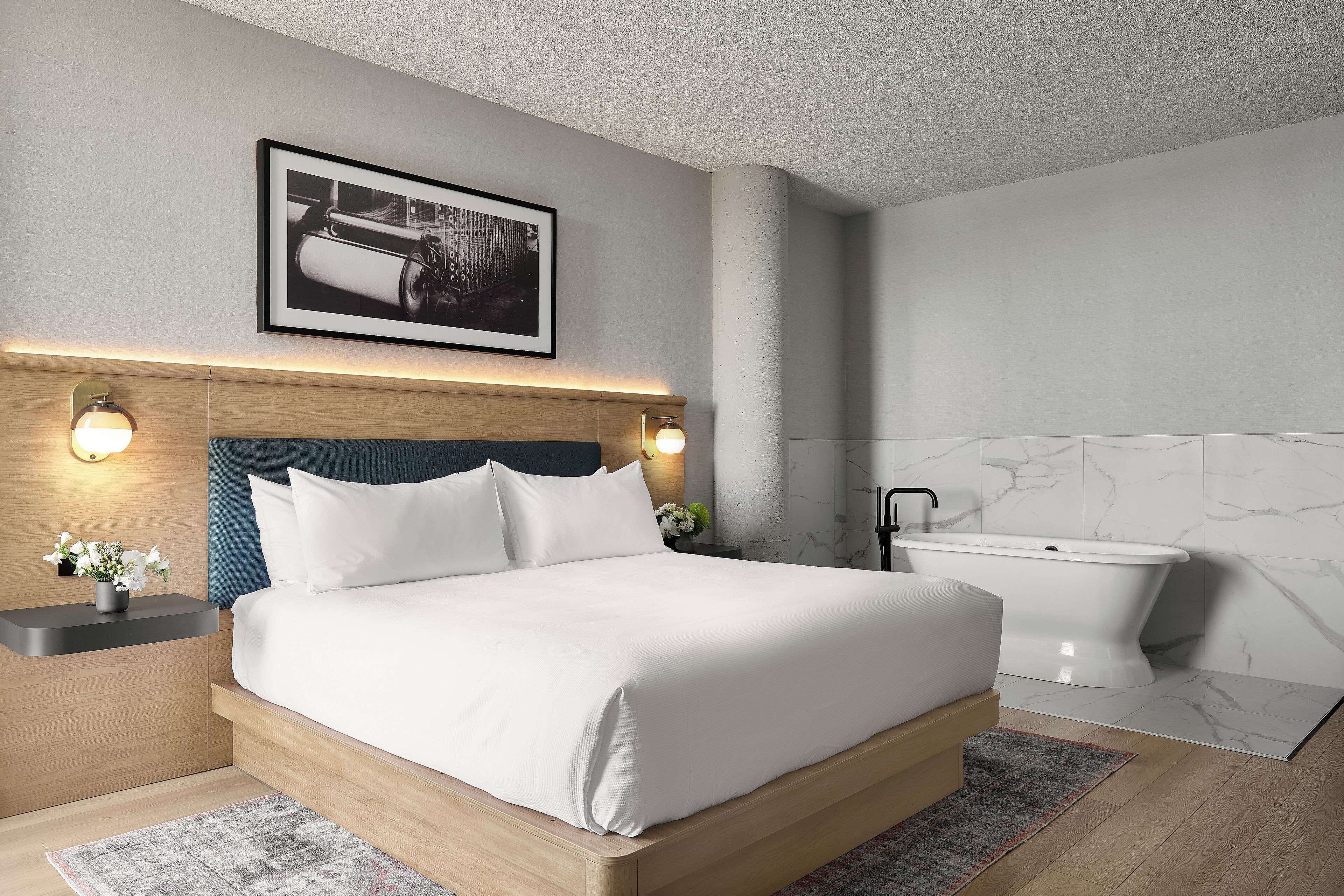 A modern bedroom with a wooden bed, bedside lamps, a framed picture, a white bathtub, and marble walls.