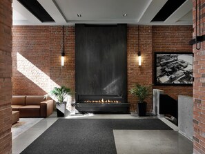 A modern living room with a fireplace, brick walls, and a large window allowing natural light. - Hôtel MOCO Valleyfield (Salaberry De Valleyfield)