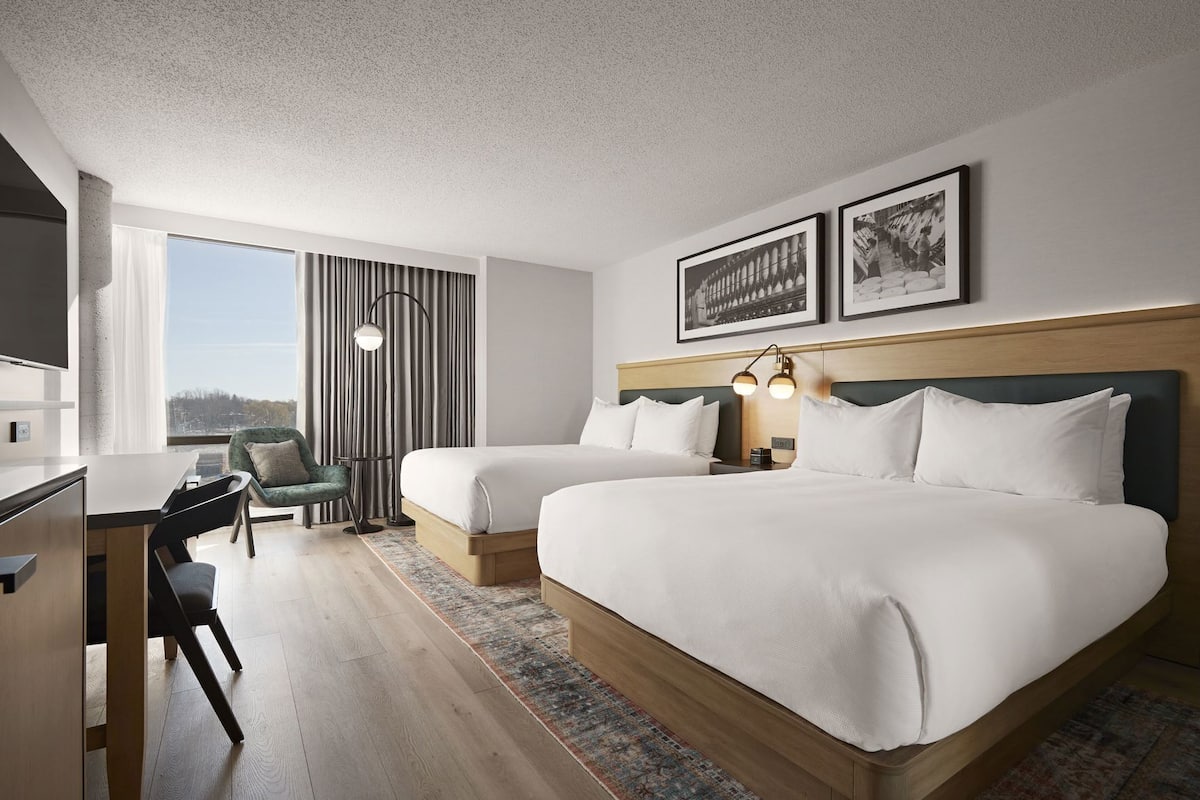 A hotel room with two beds, a desk, a chair, a large window, and a view of the city.