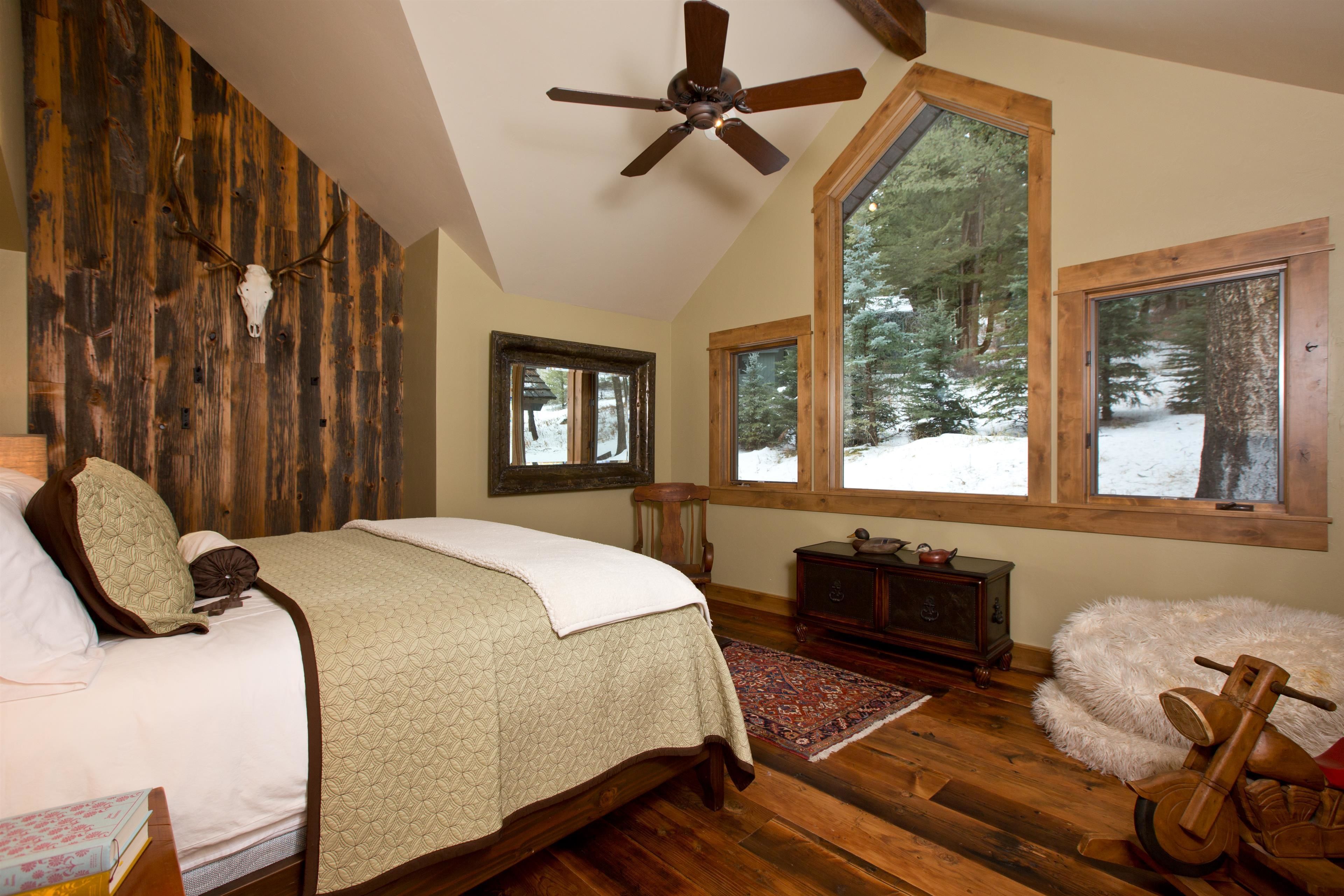 A bedroom with a wooden ceiling, a bed with a quilt, a wooden nightstand, a dresser, and a large window with a view of trees.