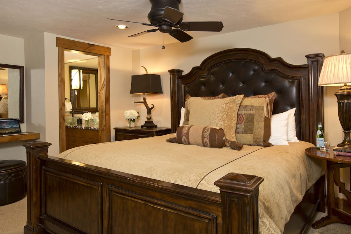 A bedroom with a large bed, a ceiling fan, a nightstand with a lamp, and a mirror.