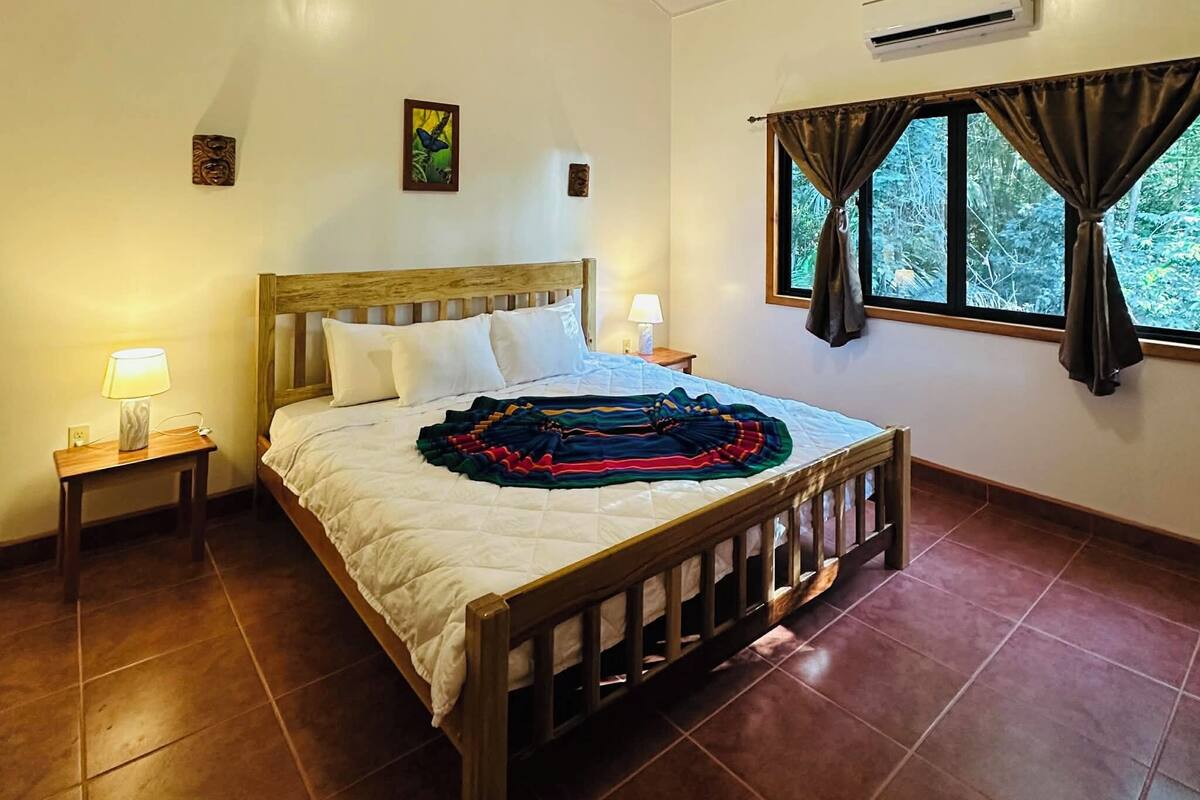 Quarto com cama de madeira, ventilador de teto, ar condicionado, janela com cortinas e mesa de cabeceira com abajur.