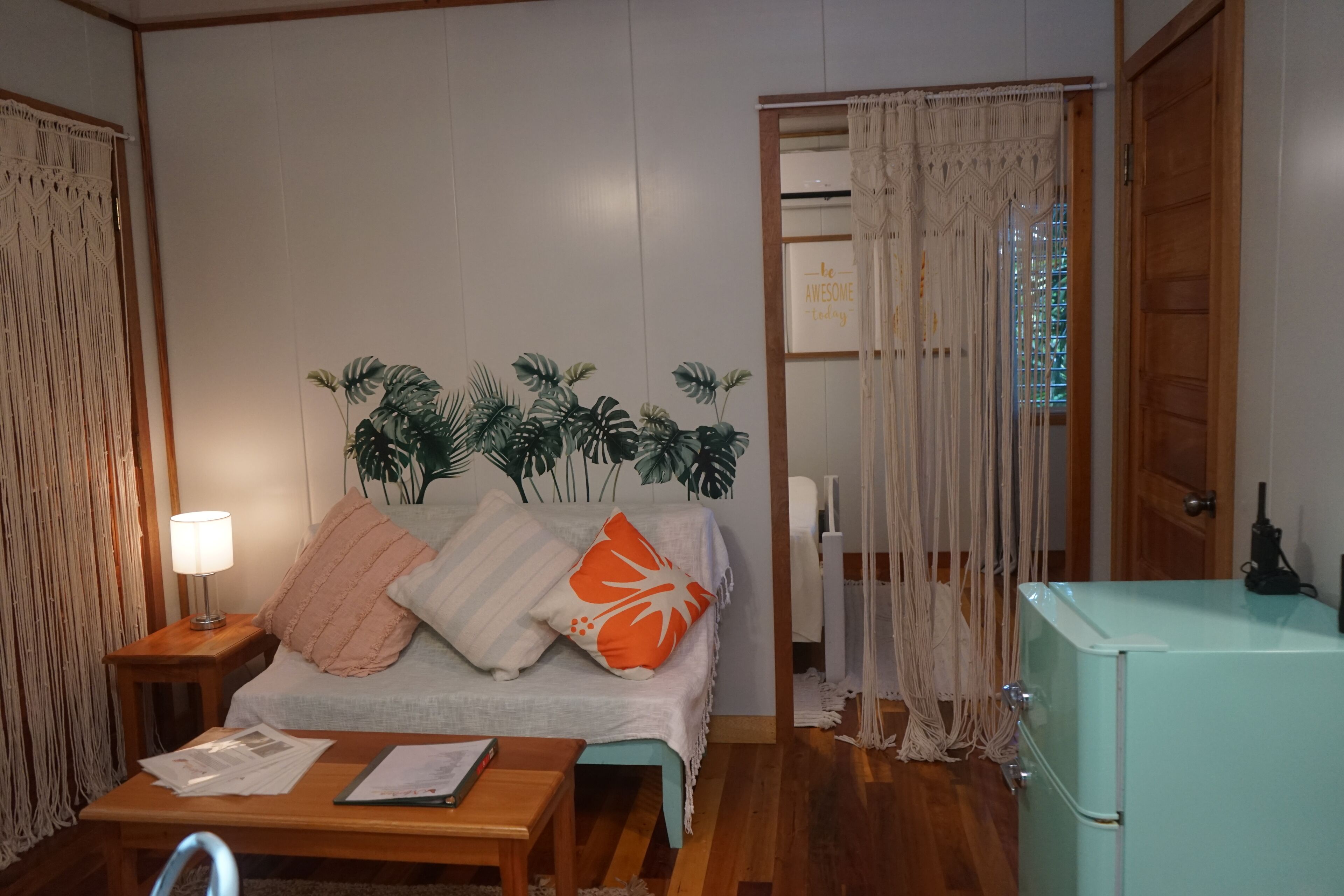 Una habitación con cama, una mesa de madera, una lámpara, un cortinero de macramé y un refrigerador antiguo.