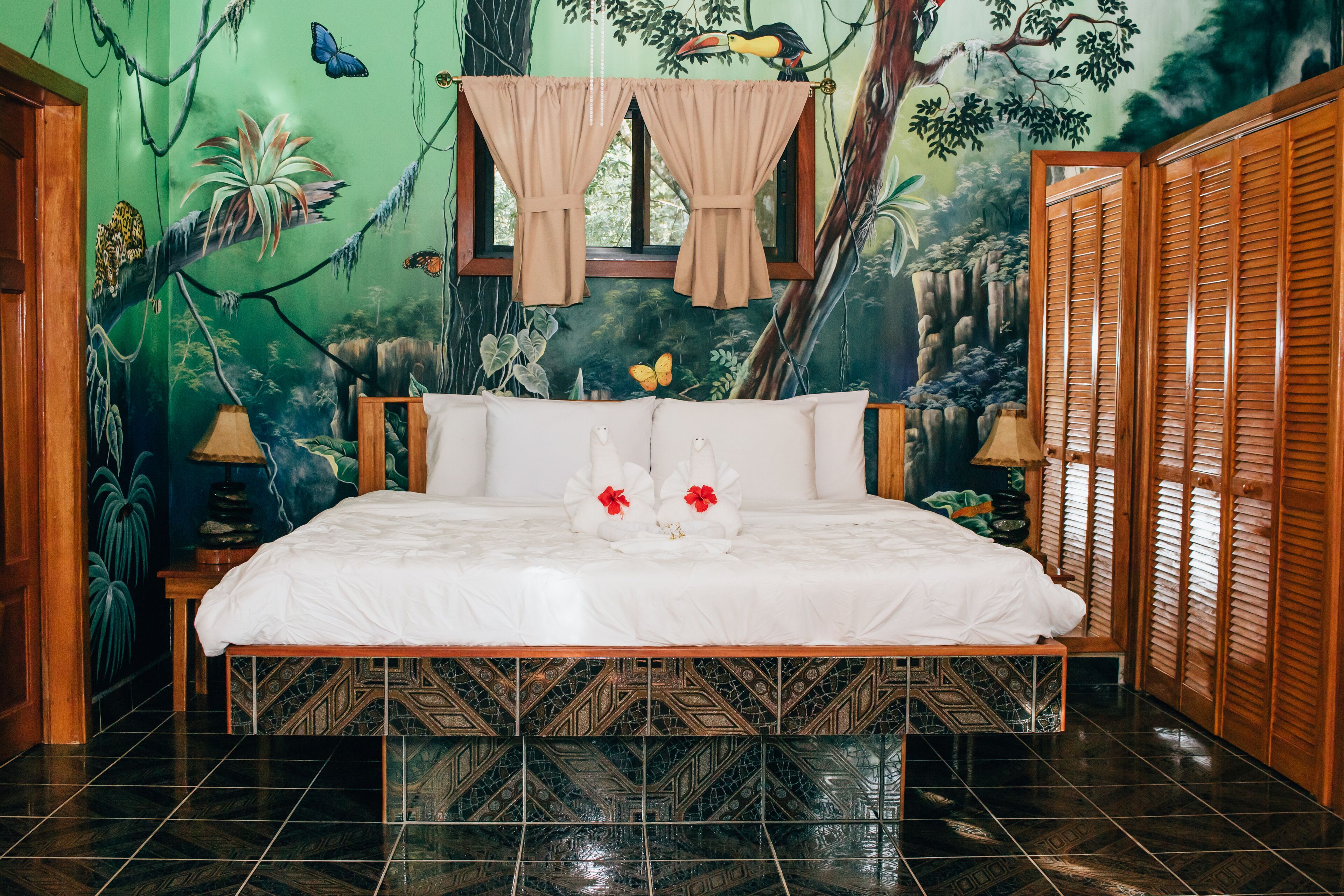 Een bed met een witte spreidek en twee rode bloemen, een raam met beige gordijnen, en een muurschildering van een boslandschap op de muur.