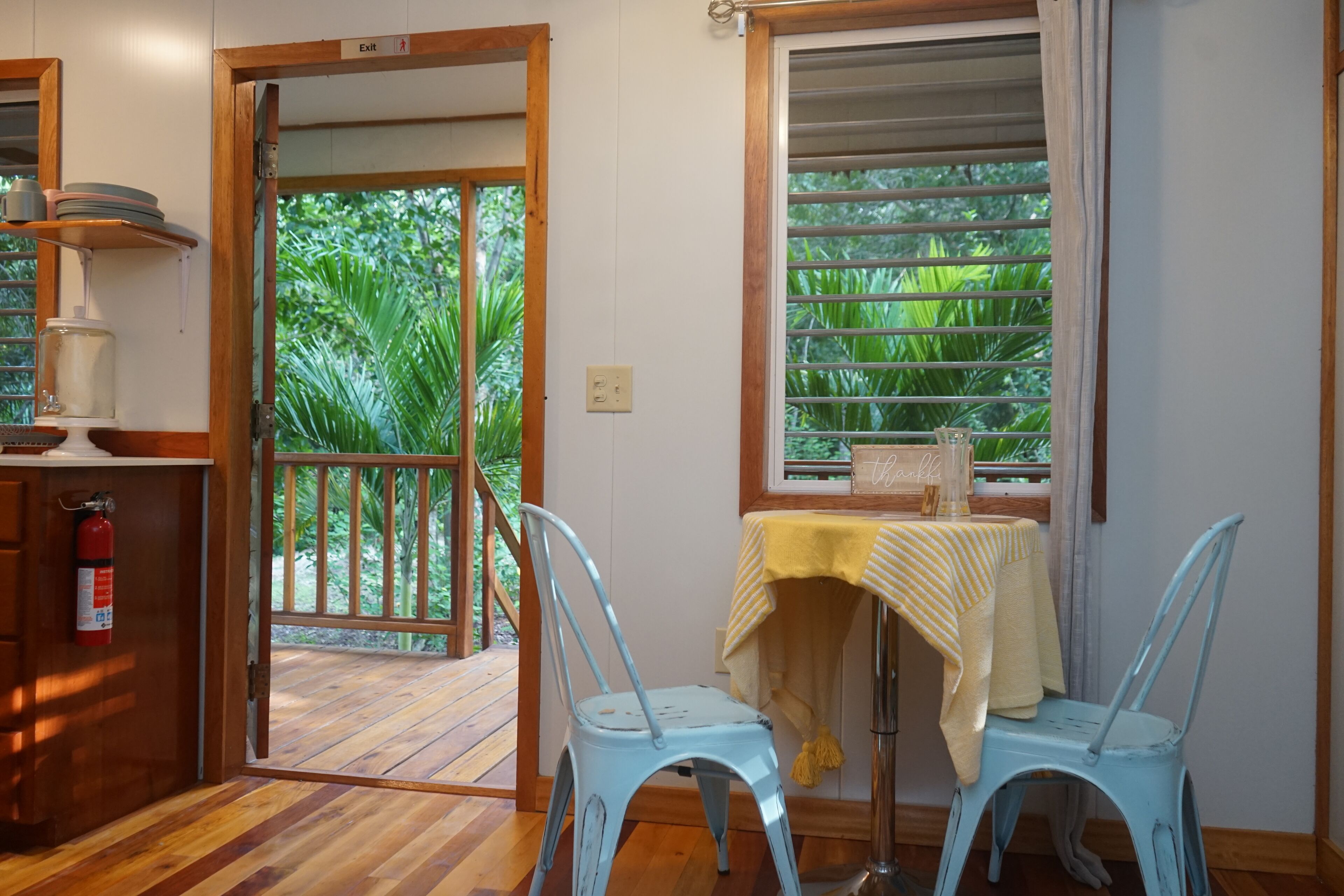 A room with a wooden floor, a table with a yellow tablecloth, two blue chairs, a window with blinds, and a door leading outside.