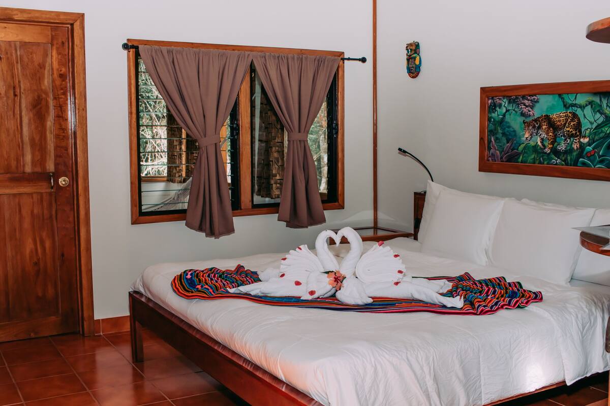 Cama bem arrumada com lençóis brancos e um edredom colorido, uma porta de madeira e uma janela com cortinas.