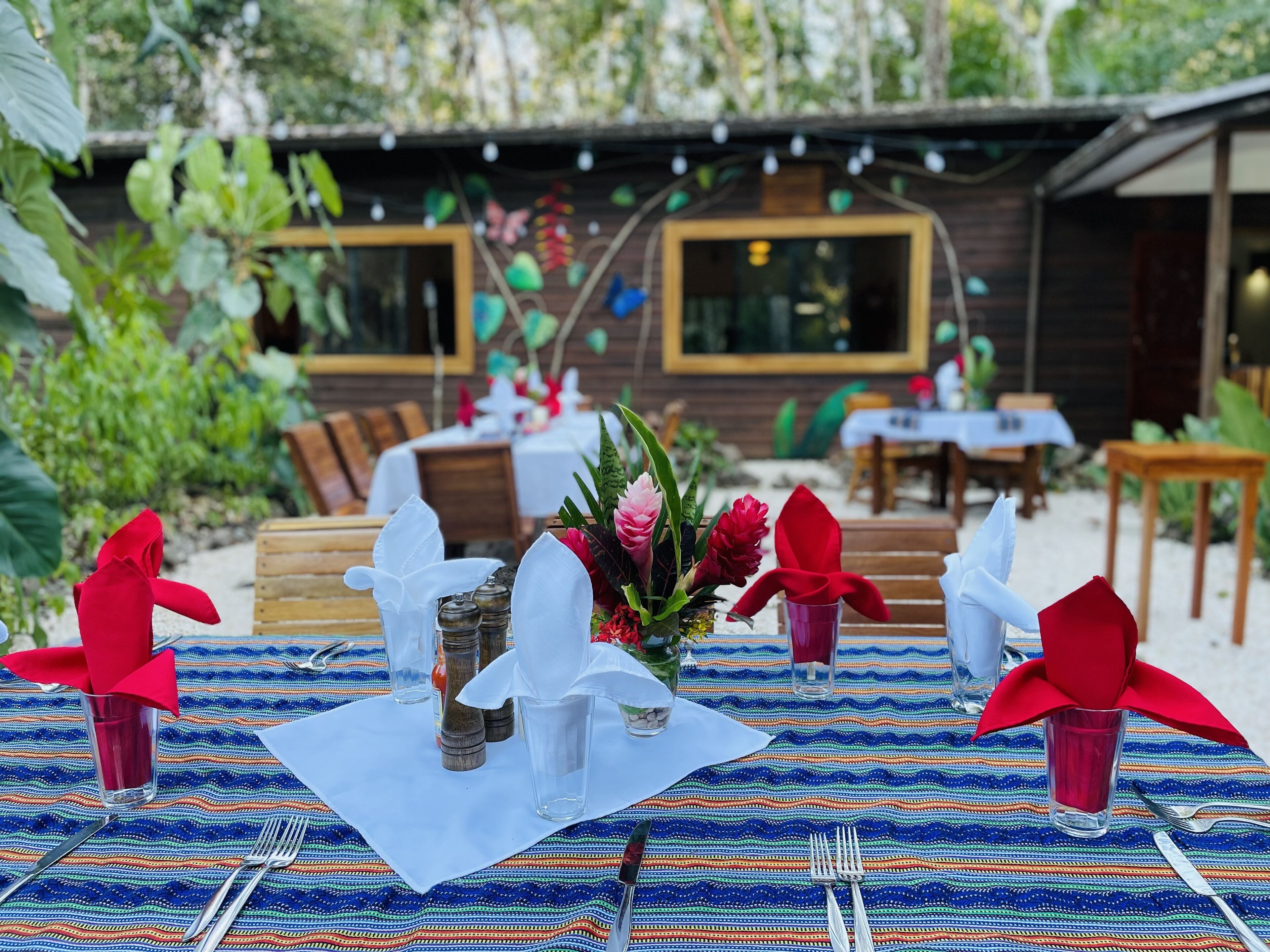 An outdoor dining area with tables set for two, featuring a colorful woven tablecloth, neatly arranged cutlery, and a centerpiece of flowers and greenery.
