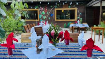 An outdoor dining area with tables set for two, featuring a colorful woven tablecloth, neatly arranged cutlery, and a centerpiece of flowers and greenery.