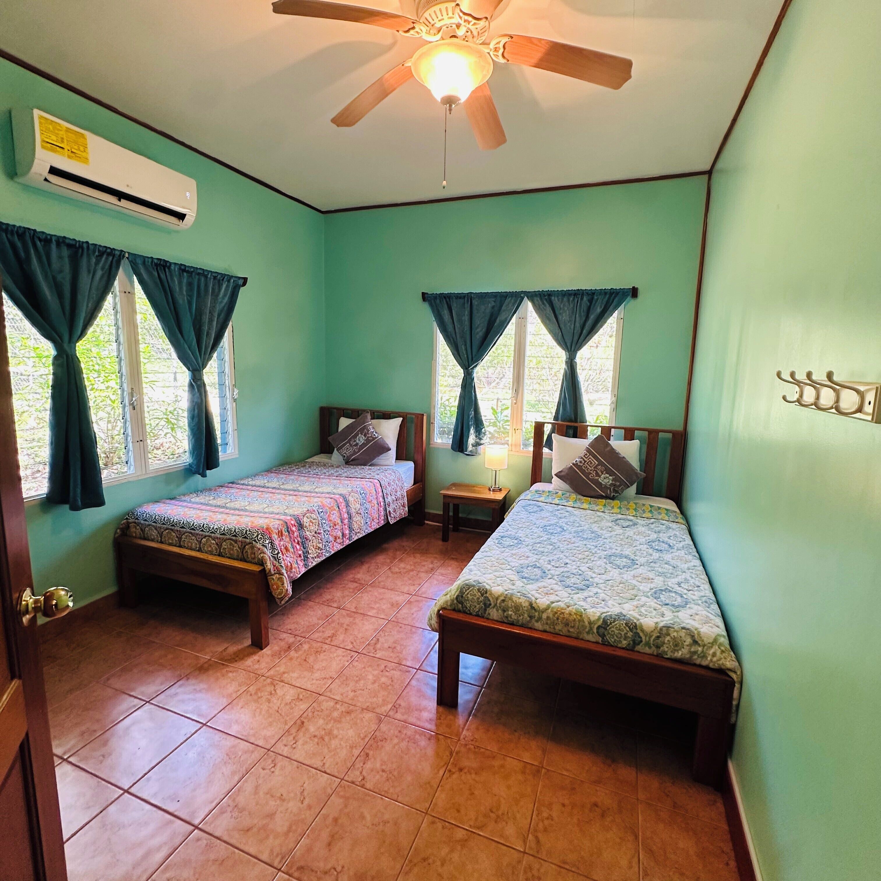 A room with two wooden beds, an air conditioning unit, a ceiling fan, and a small table with a lamp.