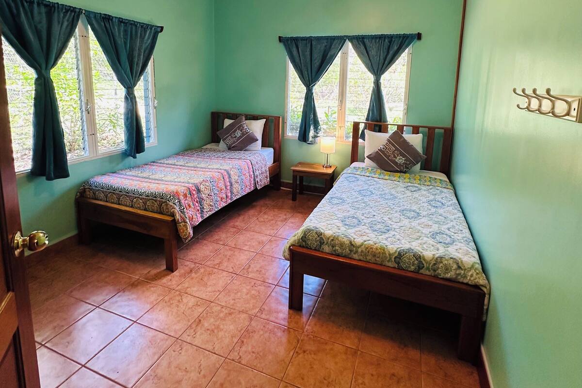 Quarto com duas camas de madeira, um ar condicionado, um ventilador de teto e uma pequena mesa com luminária.