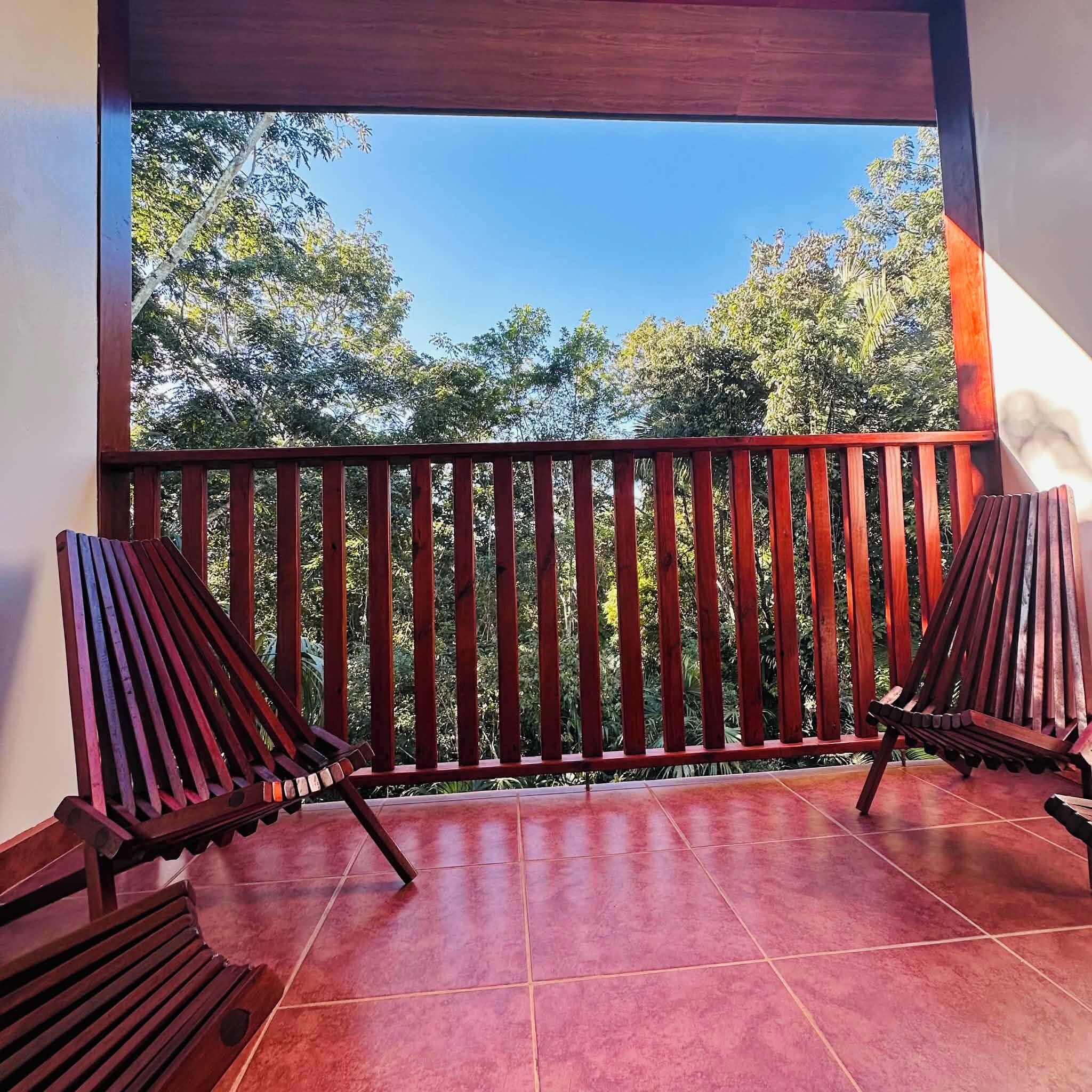 Ein Balkon mit zwei Holzstühlen, Fliesenboden und Blick auf Bäume und Himmel.